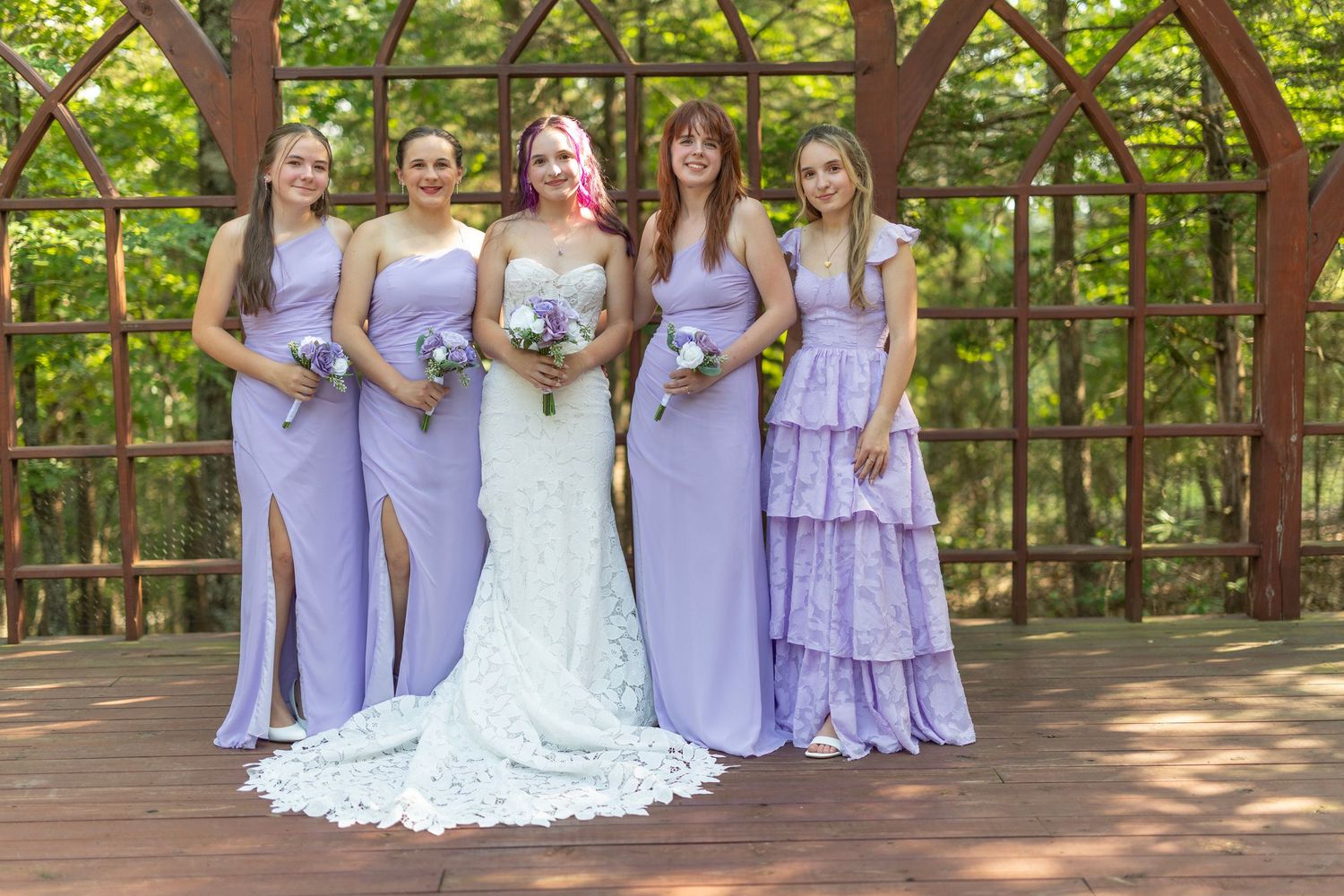 Bride in lace gown poses with three bridesmaids in lavender dresses under a wooden garden arbor.