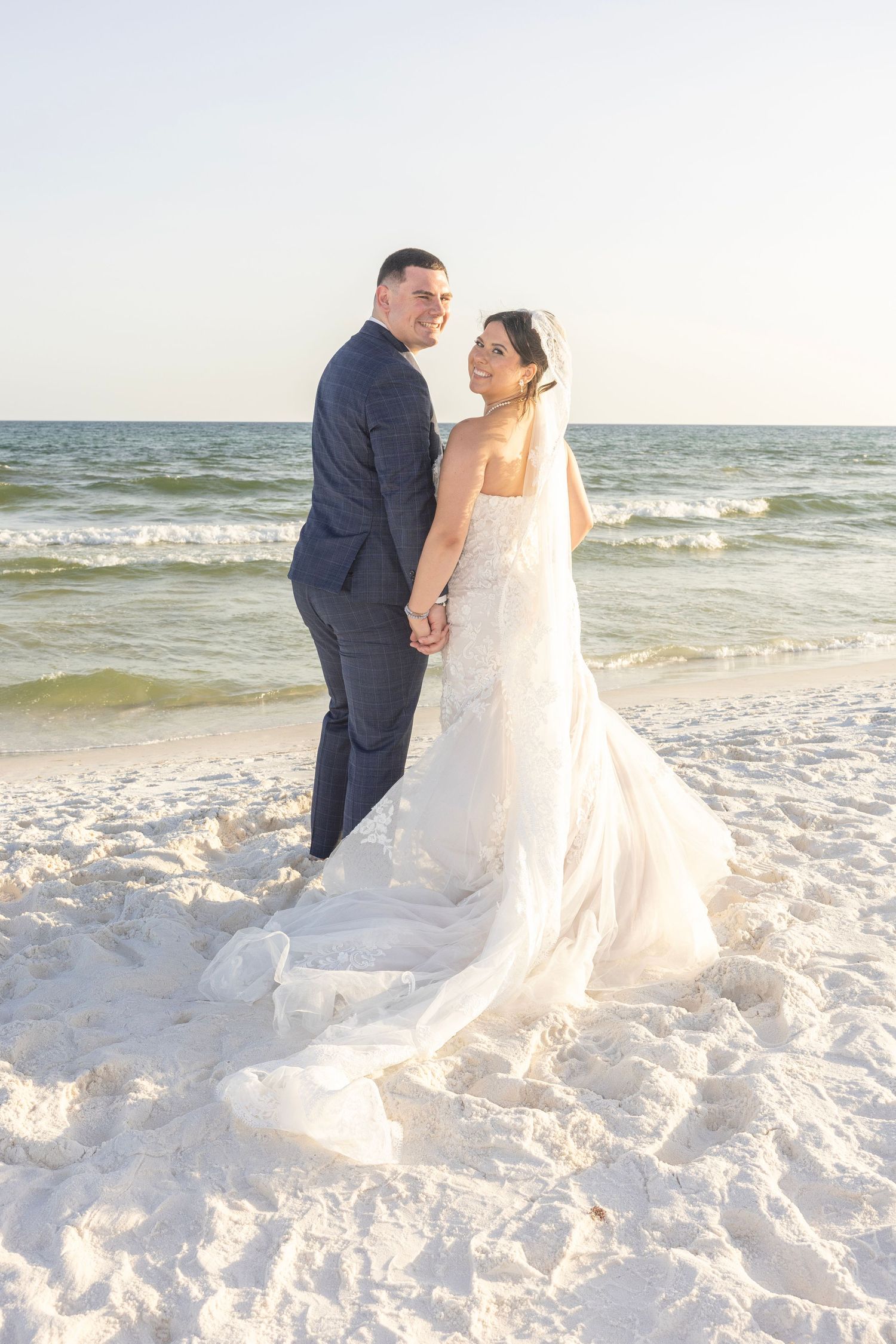 Bride and groom holding hands on a white sandy beach, with ocean waves in the background, bride wearing a flowing white gown.