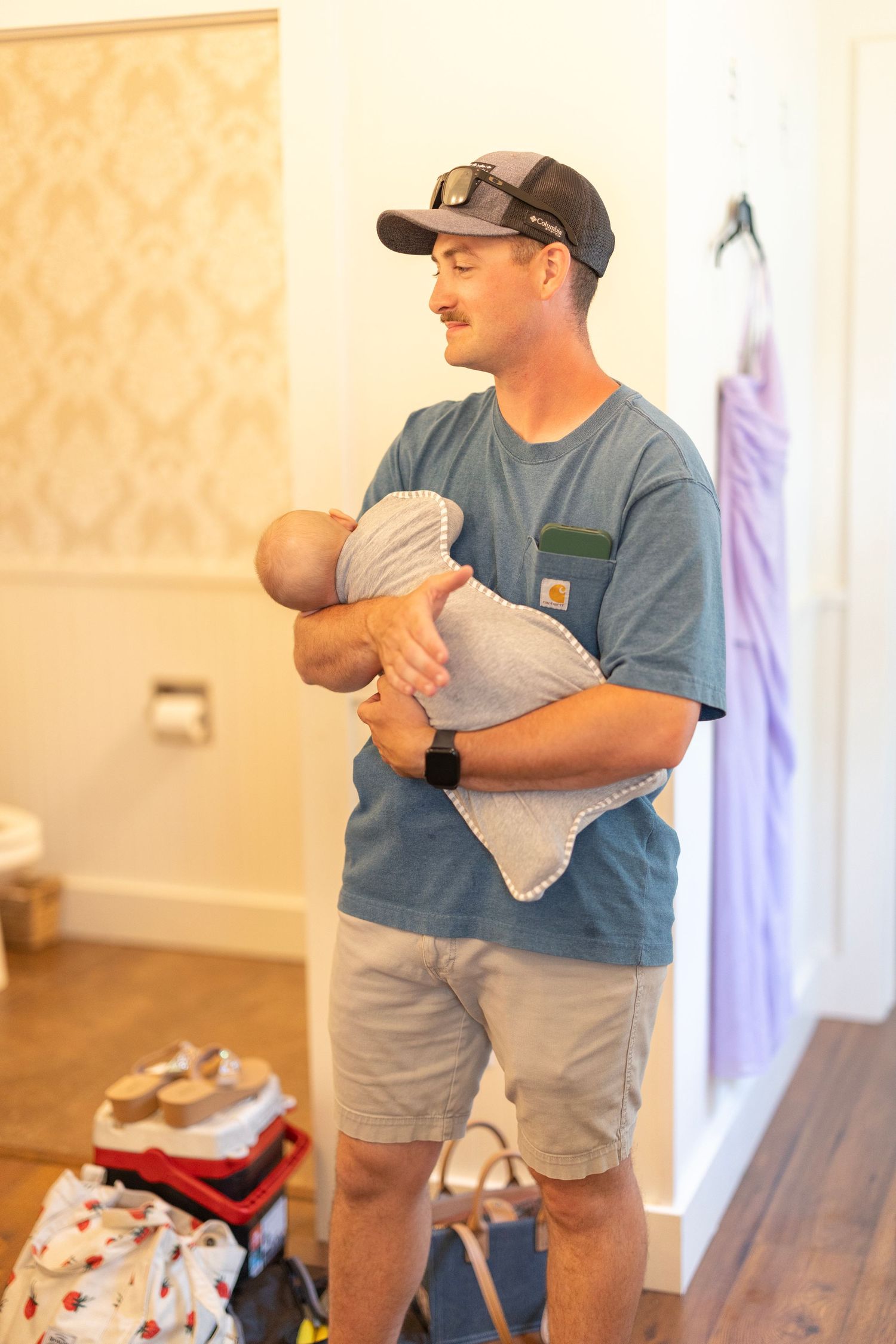 A man in a blue Carhartt t-shirt and cap gently holds a newborn baby in a cozy indoor setting.
