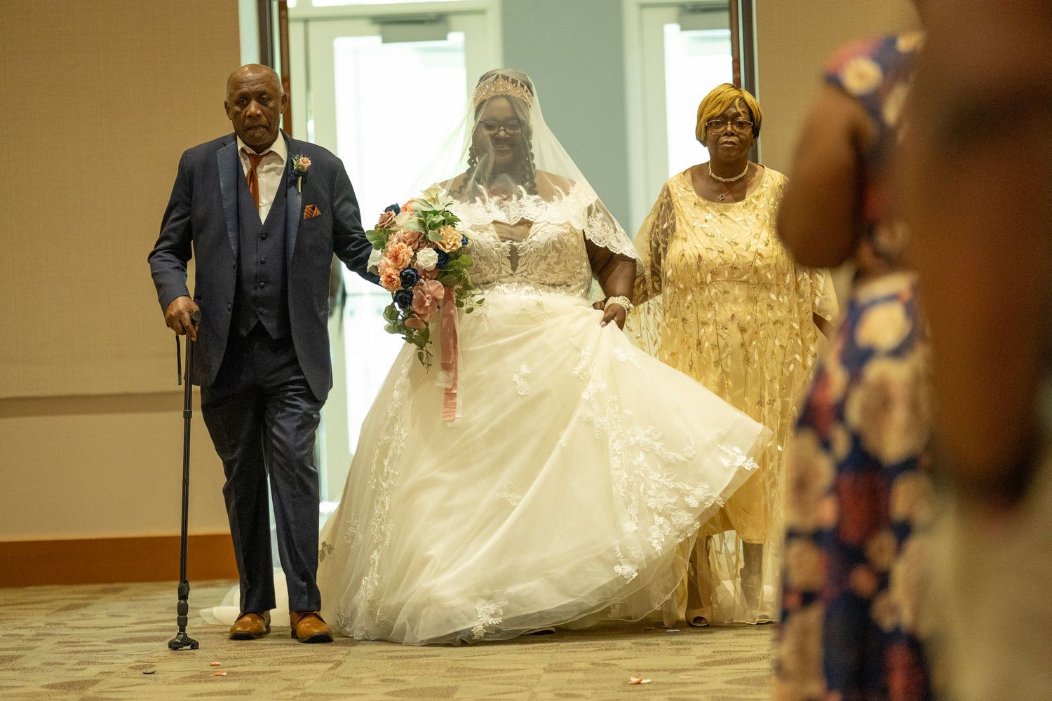 Bride in white gown walks down aisle escorted by elderly man with cane and woman in gold dress.