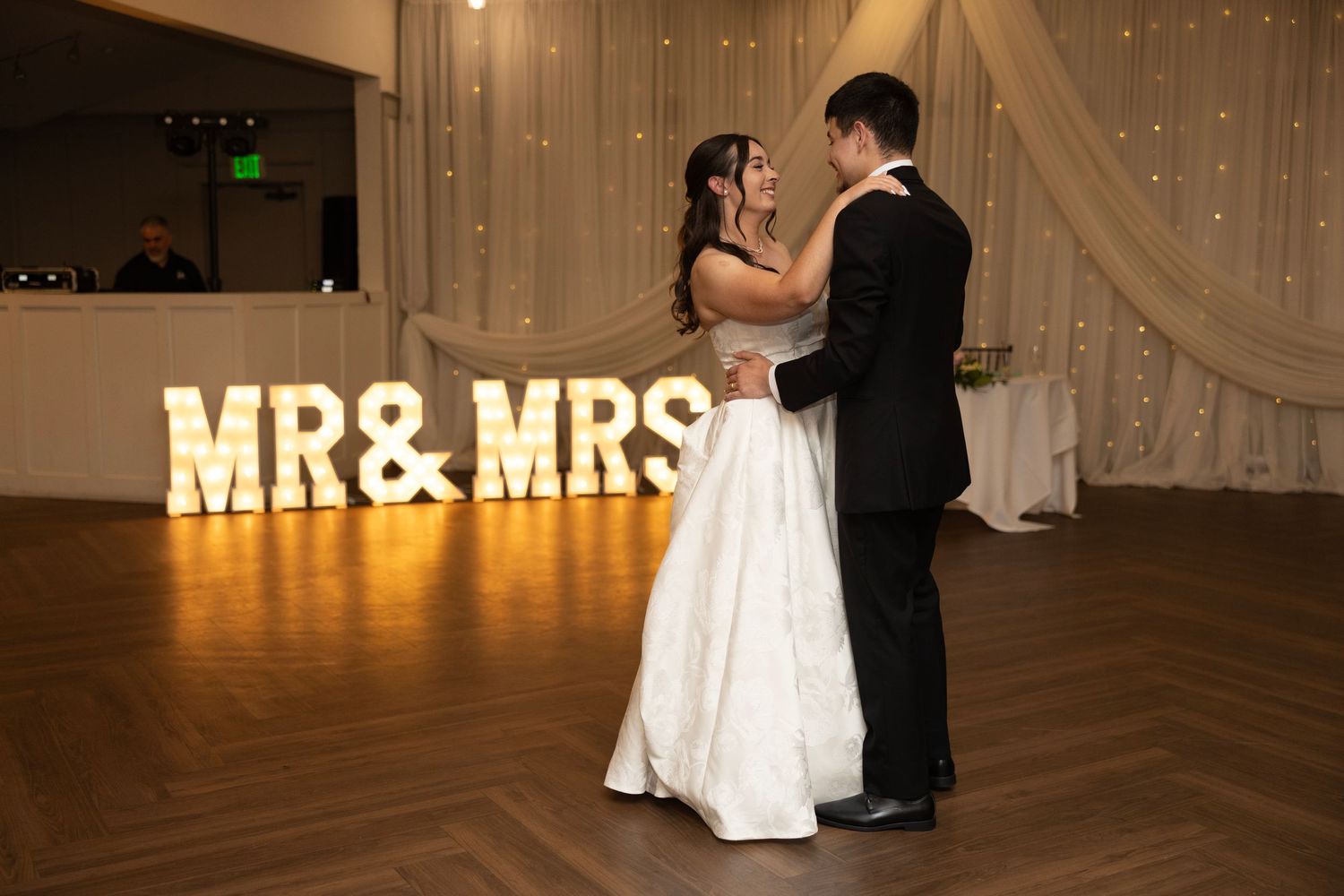 Bride and groom share first dance in front of illuminated MR & MRS marquee letters at elegant wedding reception.