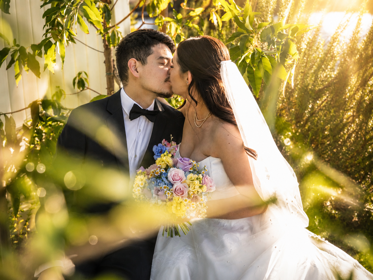 Bride and groom sharing a kiss outdoors surrounded by lush greenery with golden sunlight and a colorful bouquet.