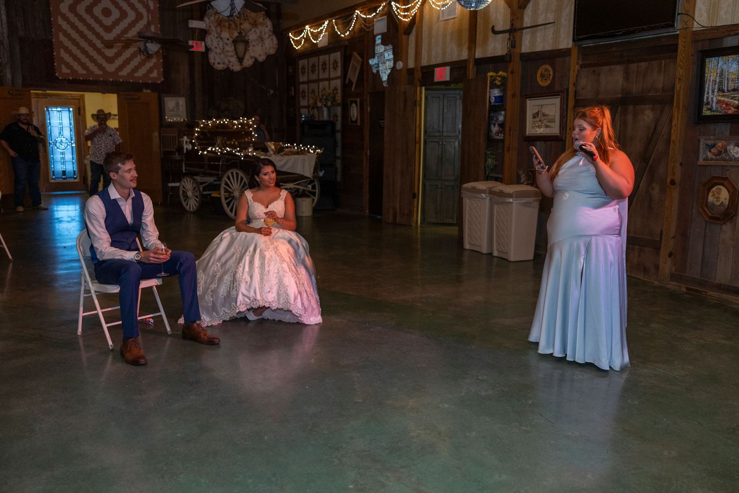 Woman in blue gown gives speech at rustic barn wedding reception while bride and groom sit listening.