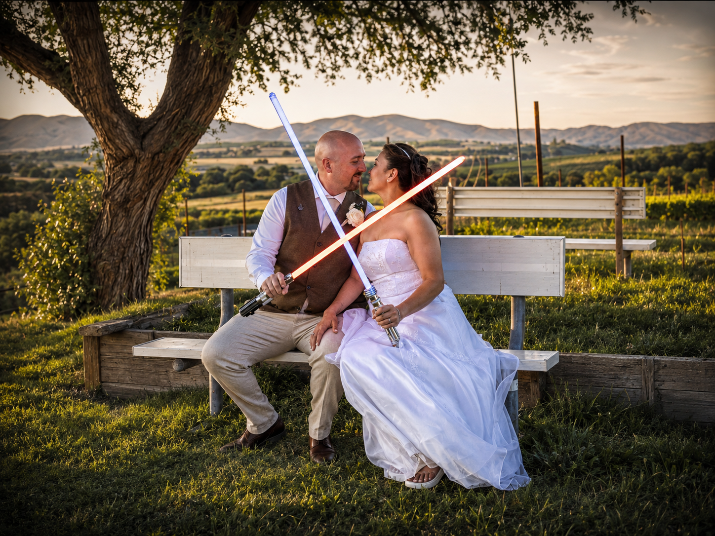 Newlywed couple kissing on bench holding lightsabers at sunset with vineyard and mountains in background.