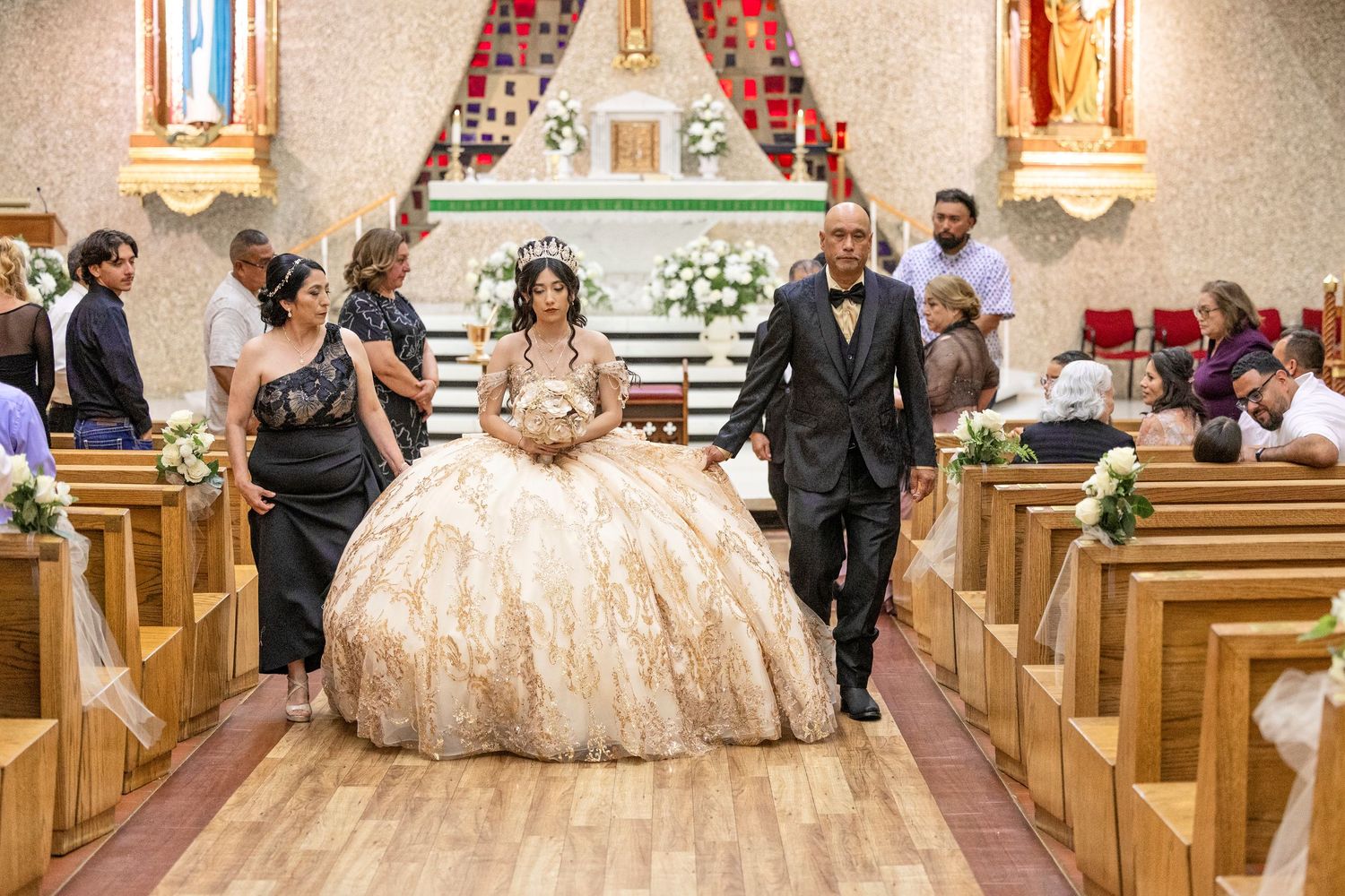 Young woman in a gold ballgown walks the church aisle during her Quinceañera ceremony with an escort.