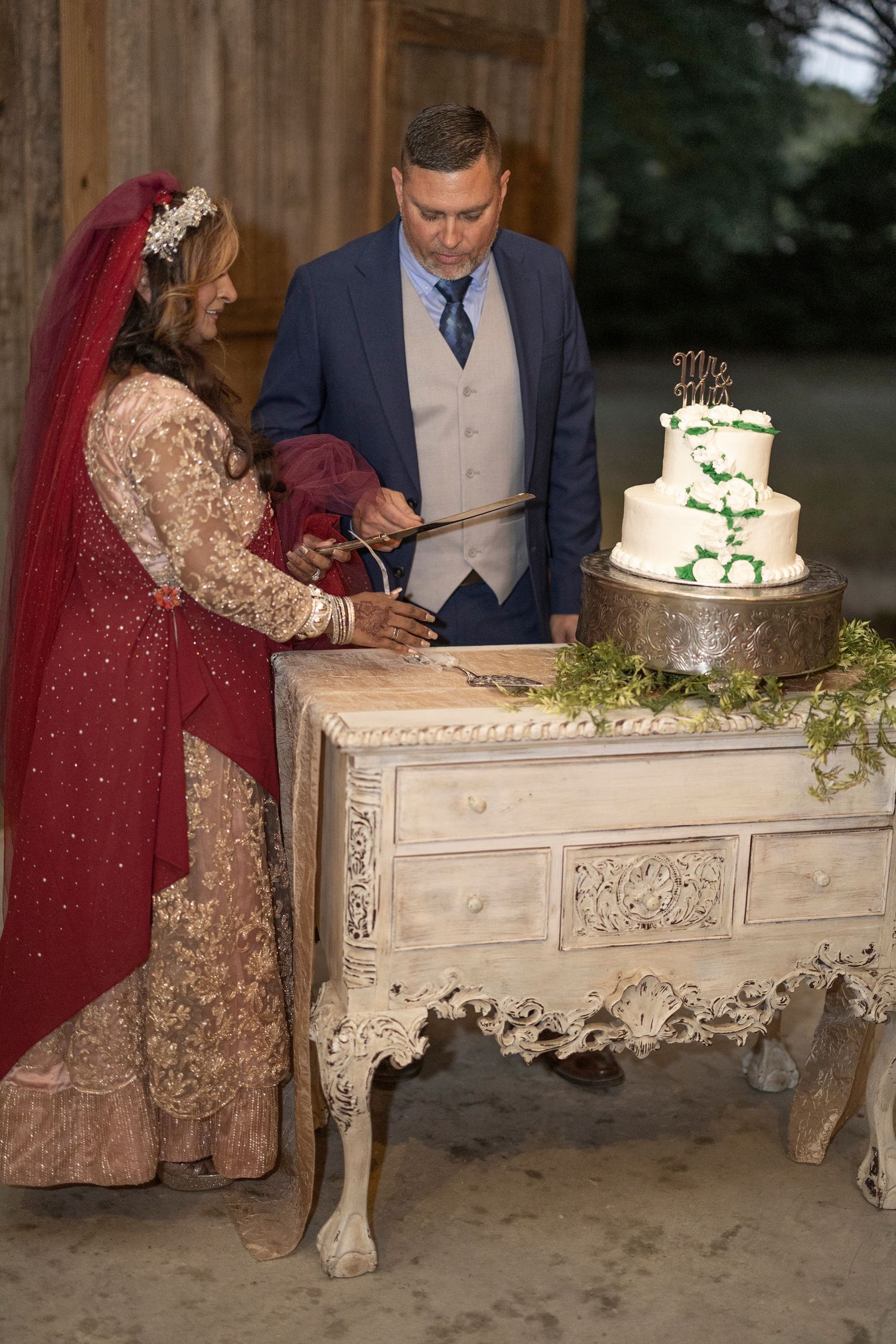 Couple cutting their elegant white wedding cake together at a rustic wooden table decorated with greenery.