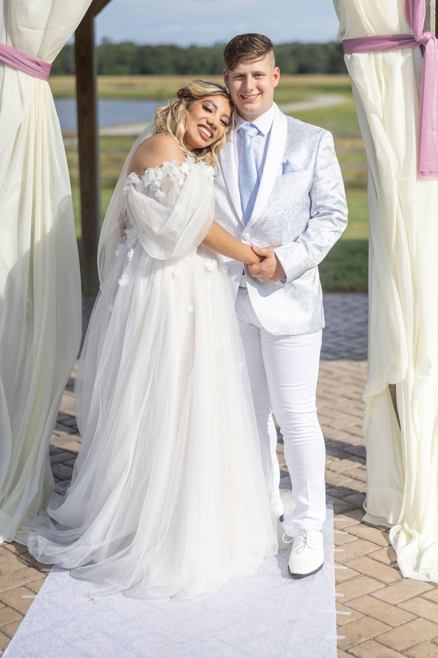 Newlywed couple in white attire pose together on brick patio by waterfront during outdoor wedding celebration.