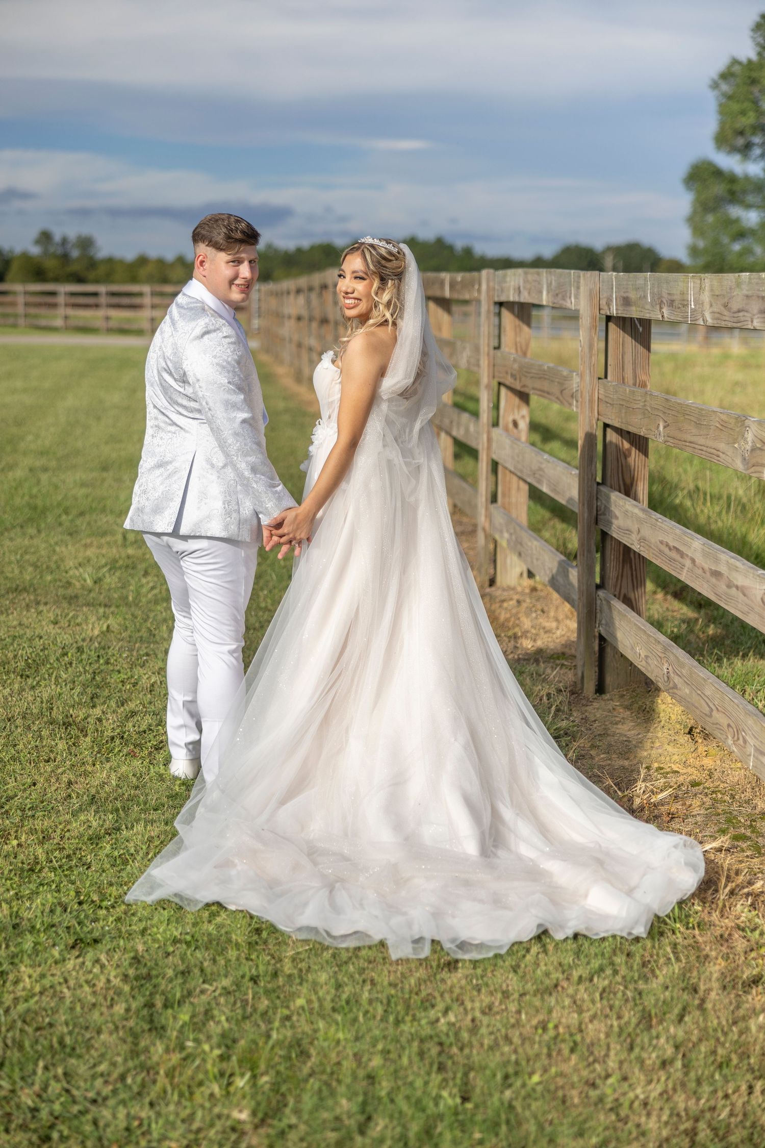 A couple in wedding attire poses romantically along a wooden fence in a grassy field on their special day.