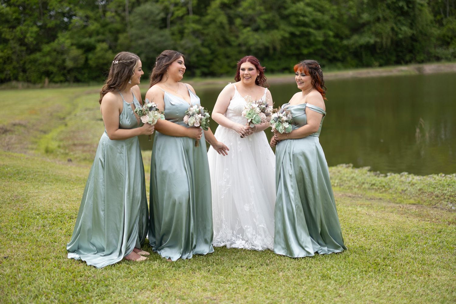 A bridal party in sage green dresses sits together on grassy lawn near a pond during an outdoor wedding celebration.
