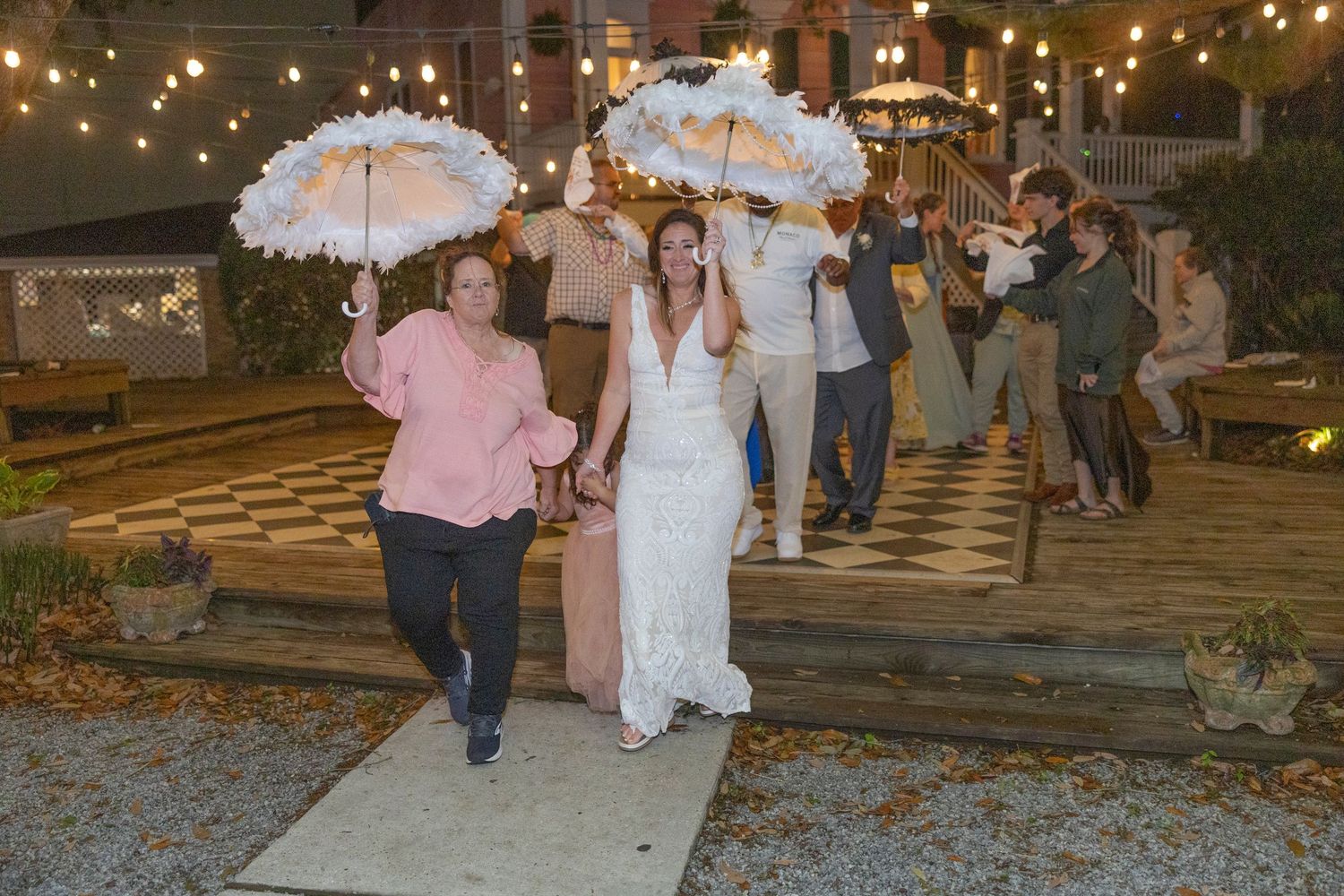 Wedding guests walk along a rustic path lit by string lights during an evening outdoor reception celebration.
