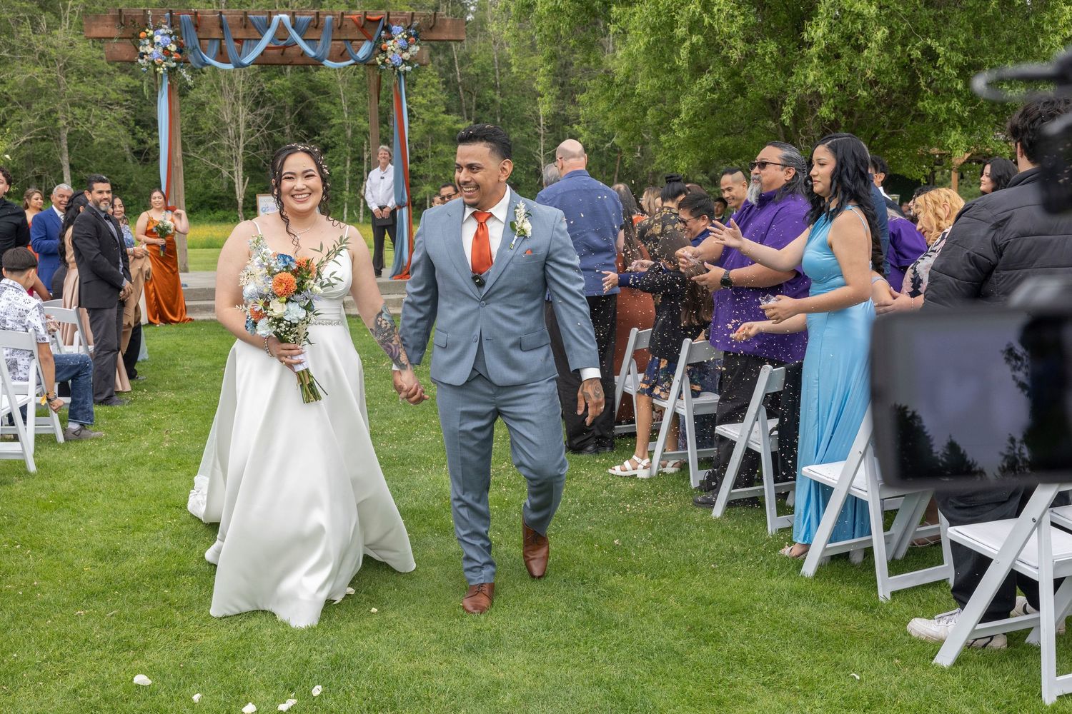 A joyful couple walks down the aisle at an outdoor wedding ceremony as guests look on from white folding chairs.