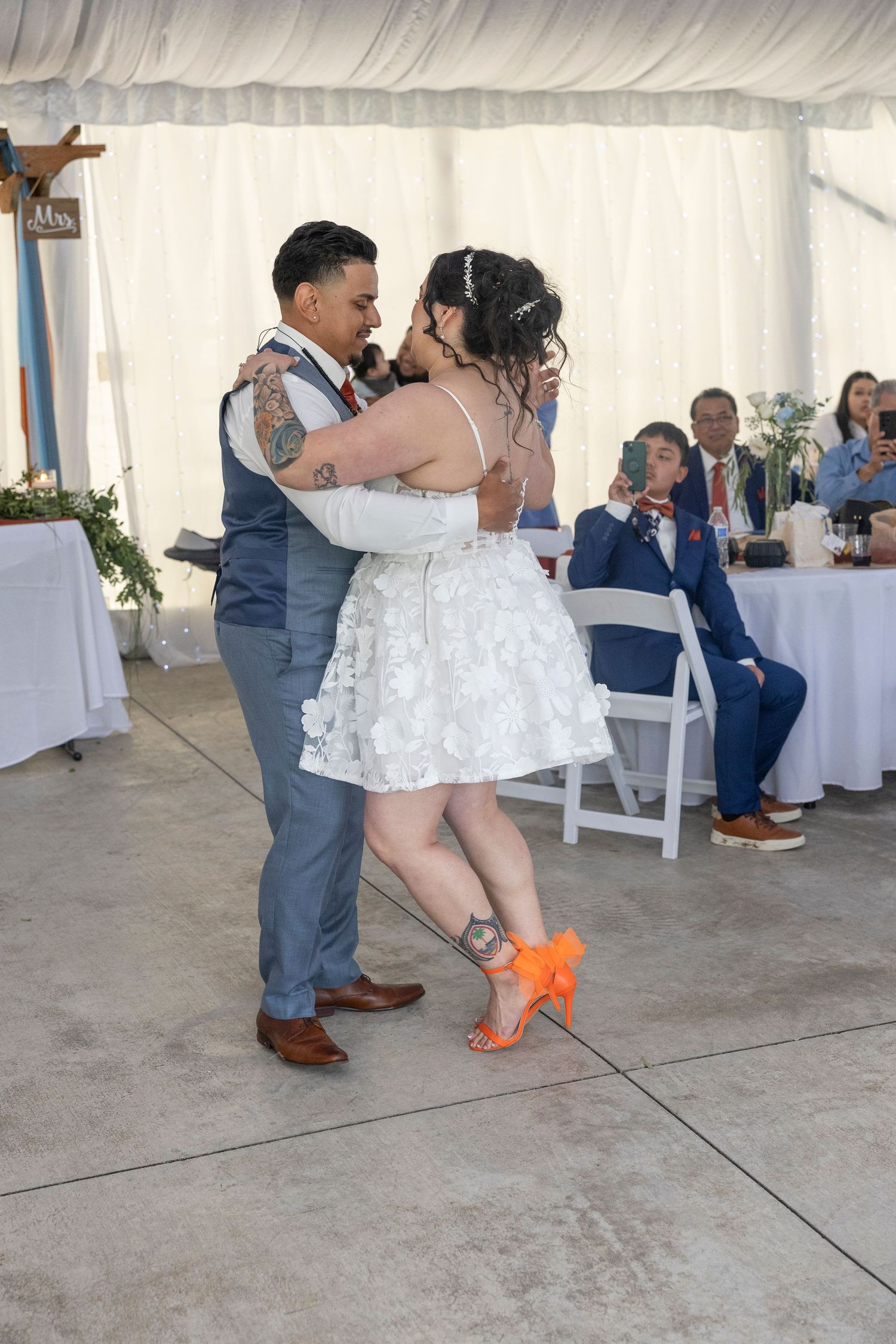 A couple shares their first dance at a wedding reception under a white tent while guests watch from their tables.
