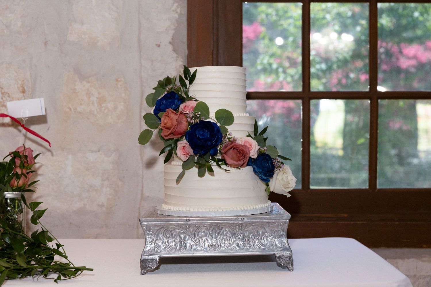 Elegant white wedding cake decorated with navy blue and coral flowers sits on silver stand by rustic window.