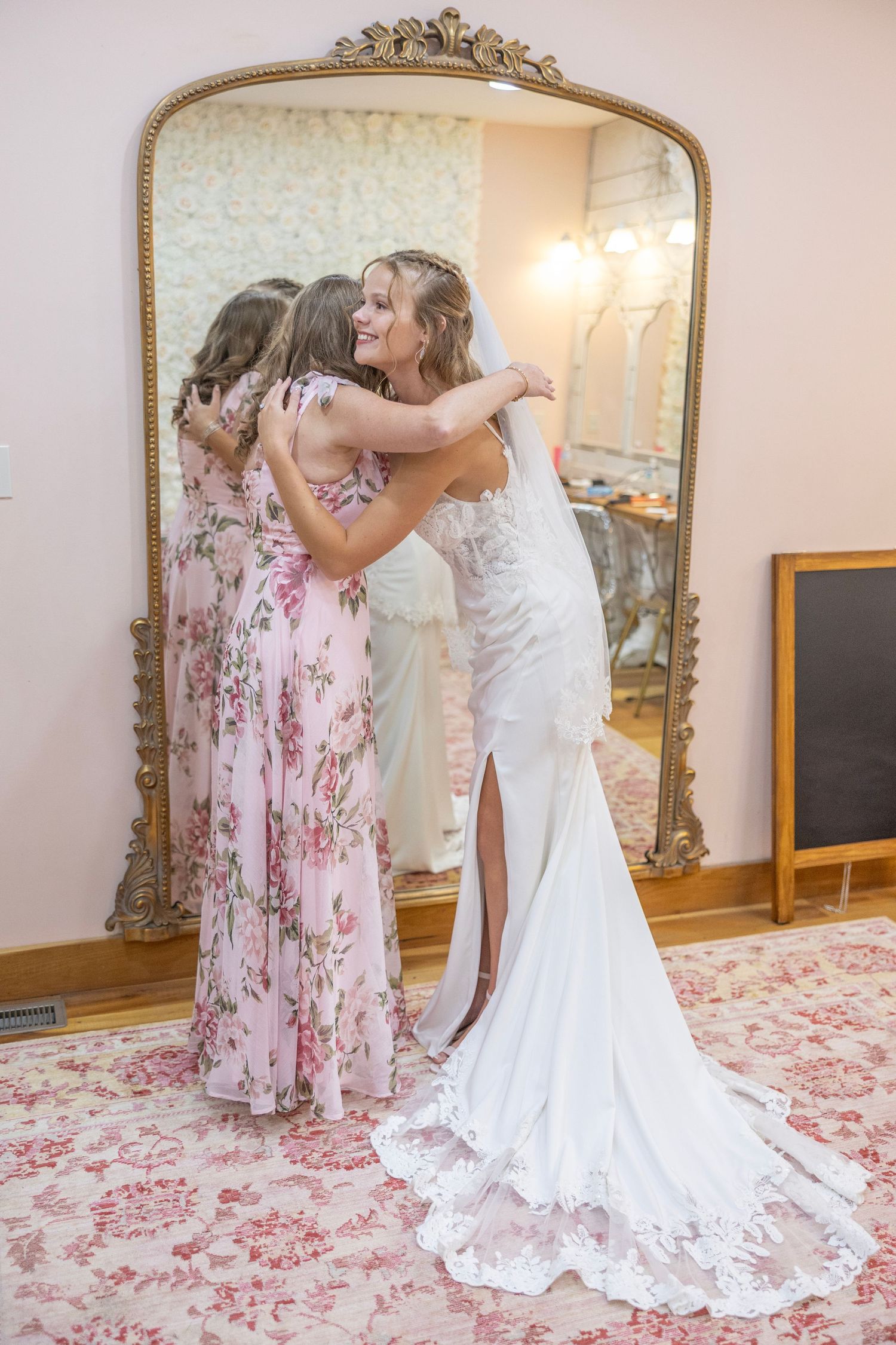 A bride in a white lace gown embraces someone wearing a pink floral dress in front of an ornate gold mirror.
