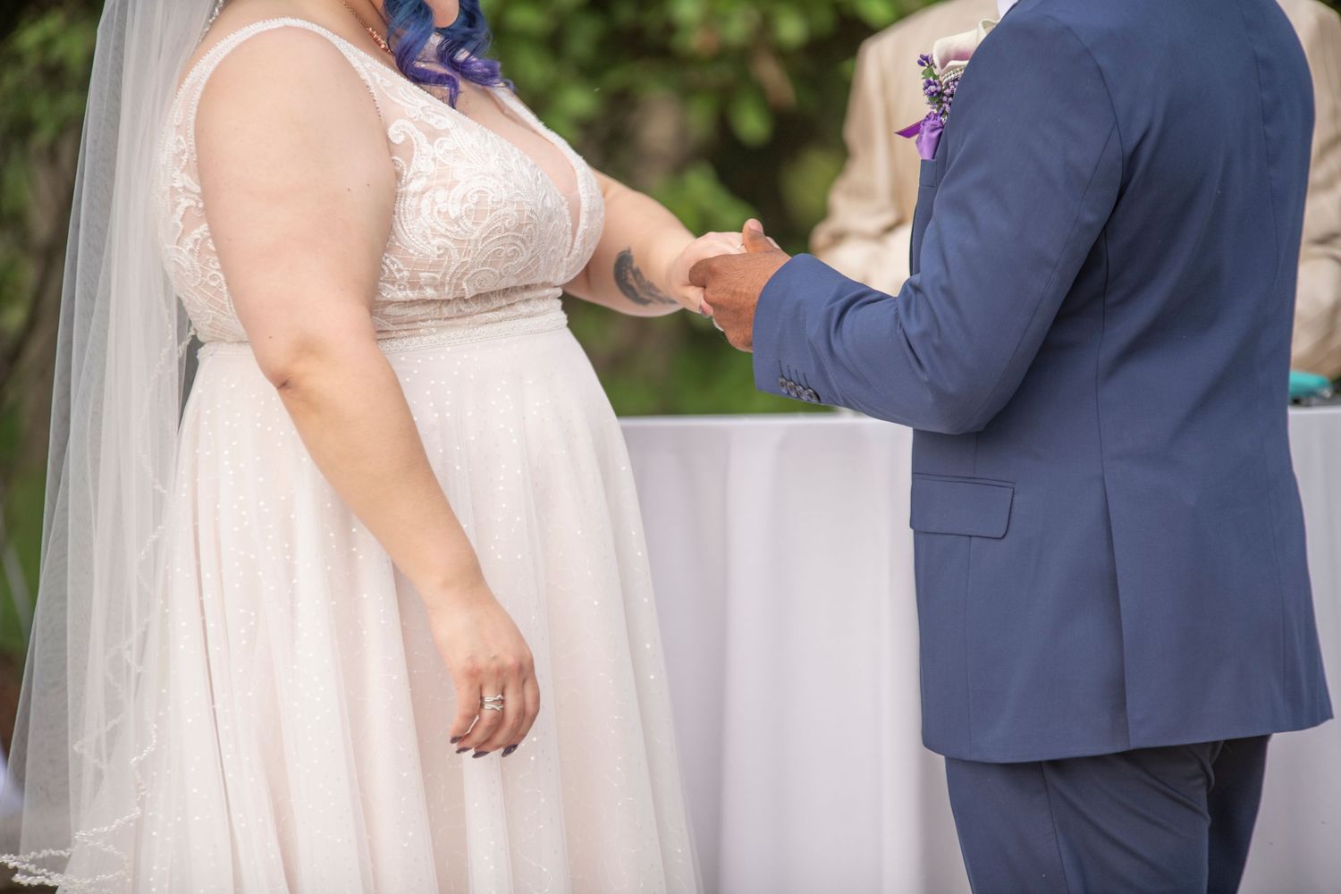 Close-up of couple exchanging wedding rings during outdoor ceremony with bride in white lace dress and groom in navy suit.