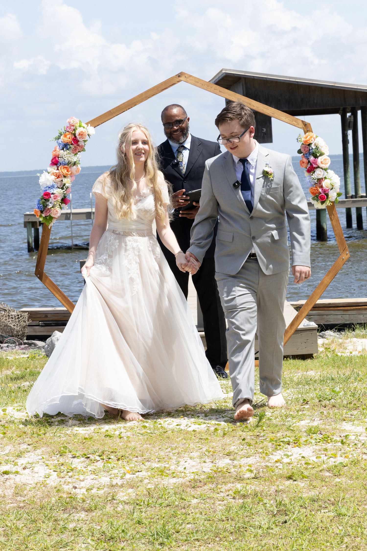 Newlyweds walk through a hexagonal floral arch at their waterfront wedding ceremony on a sunny day.