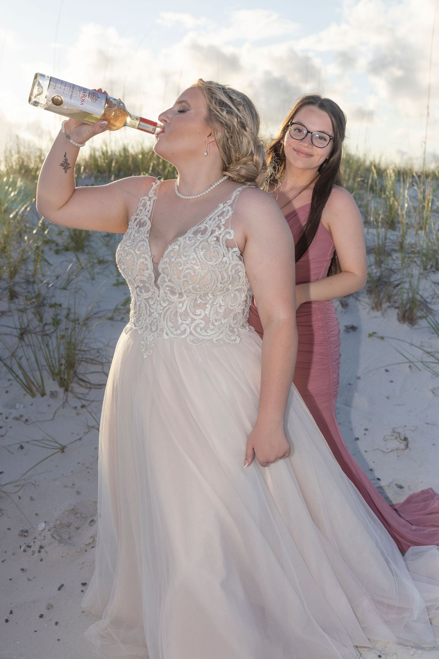 Two people in formal attire pose on a sandy beach, with one drinking from a bottle at sunset.