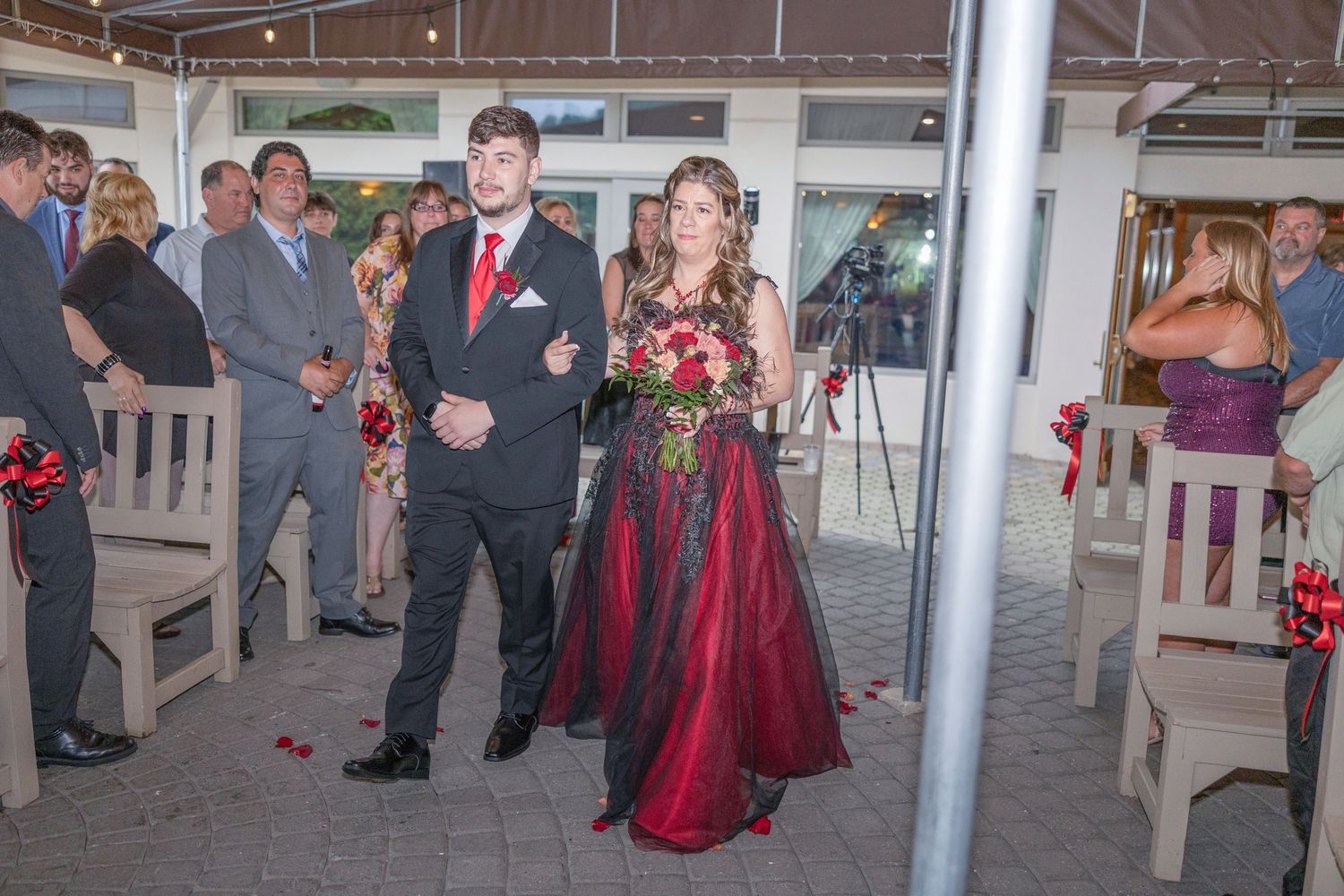 Wedding guests in formal attire gather under a covered outdoor venue with red decorative accents.