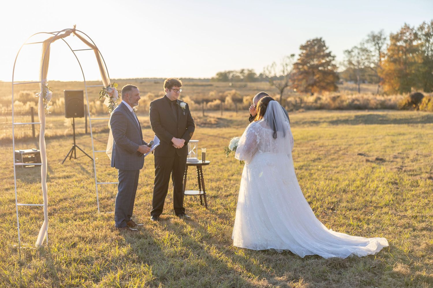 A wedding ceremony takes place in a golden field at sunset with people gathered in formal attire.