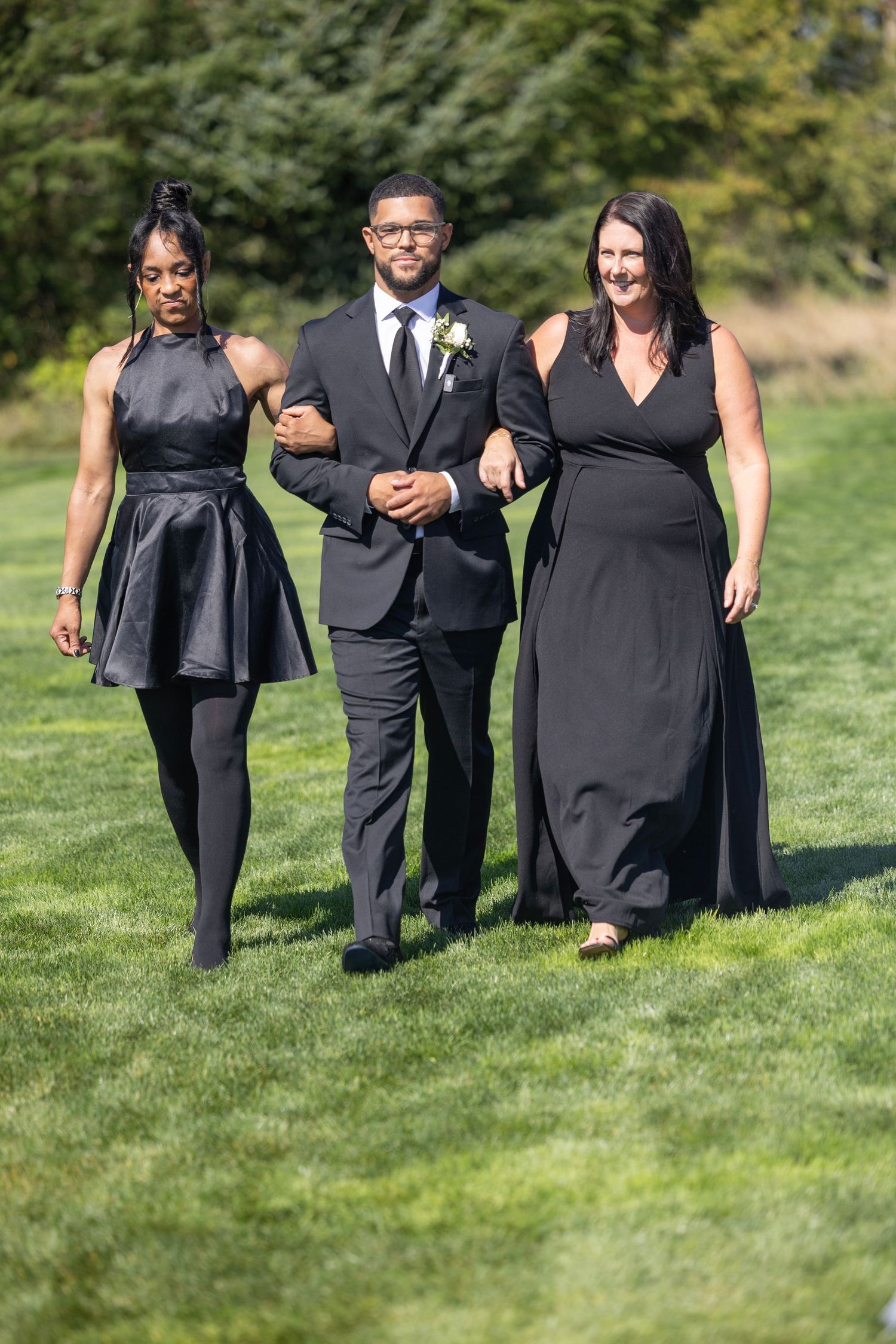Wedding party members in black formal attire walking together on a green lawn during an outdoor celebration.