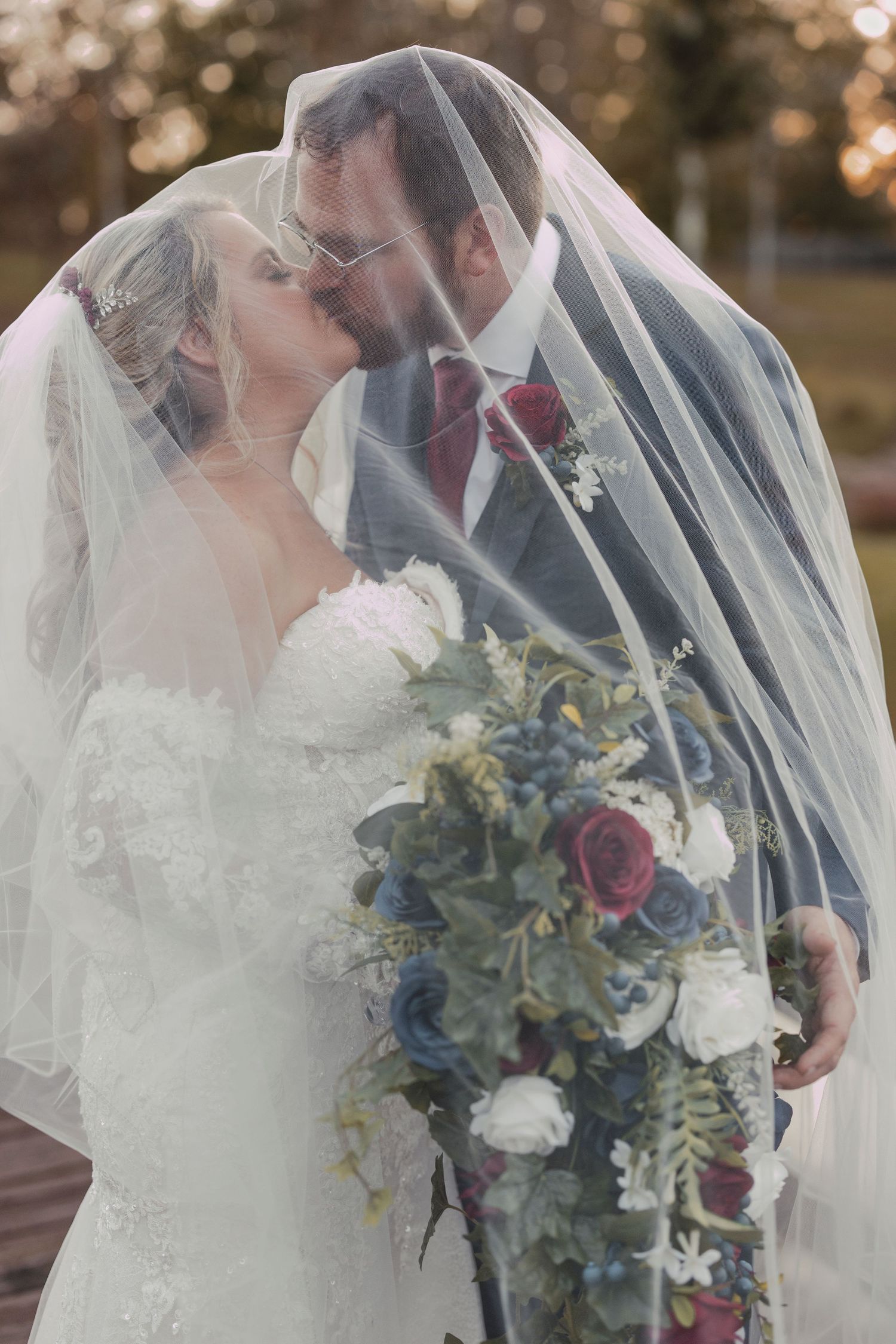 Romantic wedding couple shares intimate moment under veil with blue and burgundy floral bouquet at outdoor ceremony.