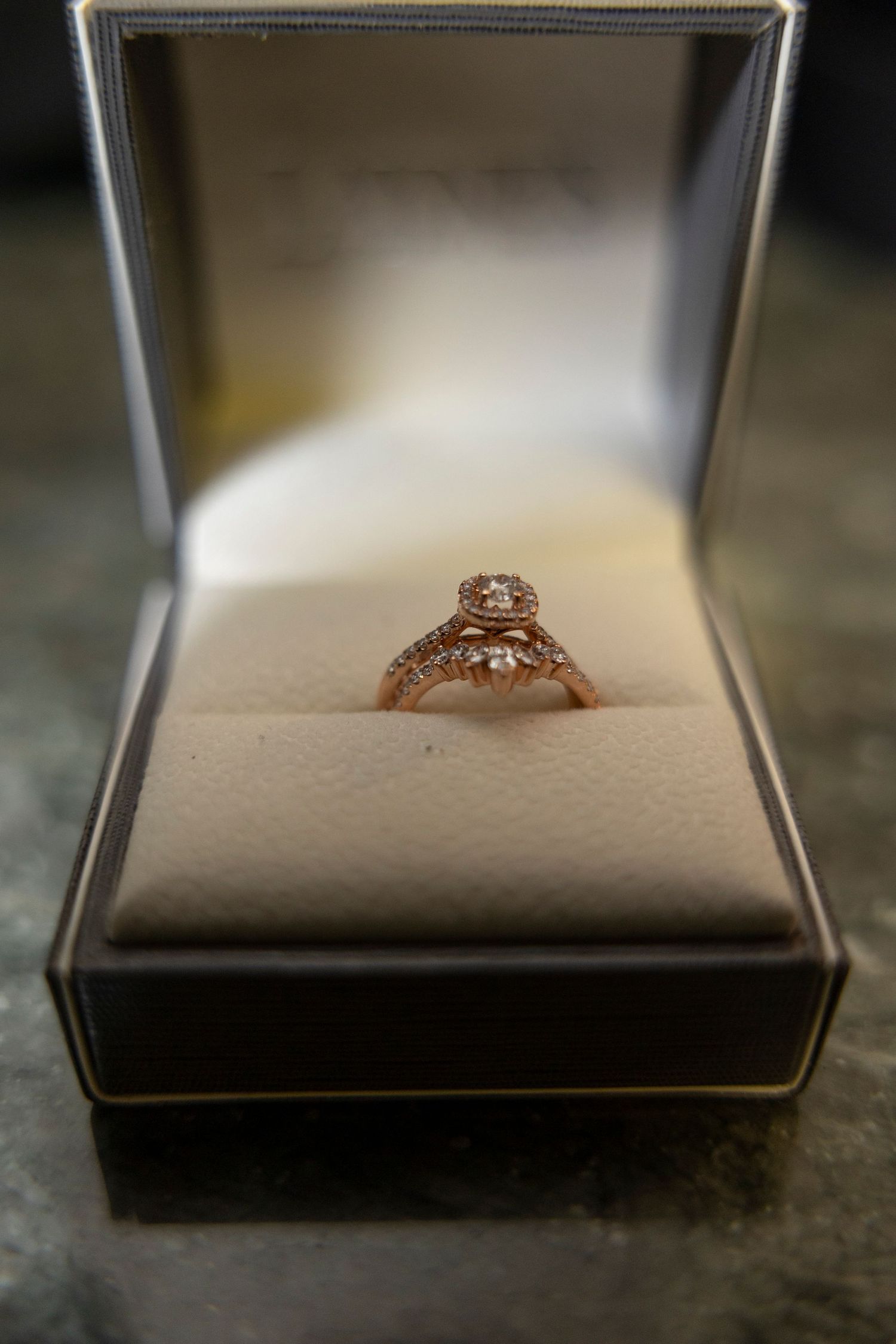 Rose gold engagement ring with intricate details sits in an open grey jewelry box.