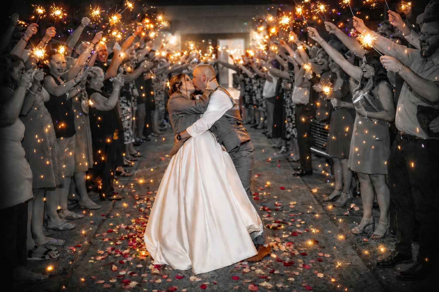 Bride and groom share a romantic kiss under a sparkler tunnel surrounded by wedding guests holding sparklers.