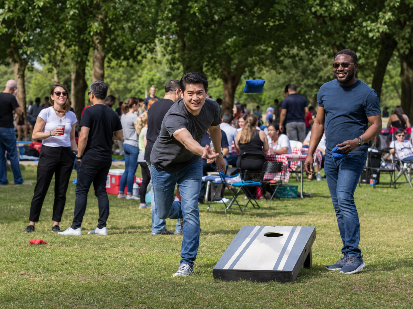 Two men playing cornhole on grass at an outdoor park gathering with people socializing in the background.