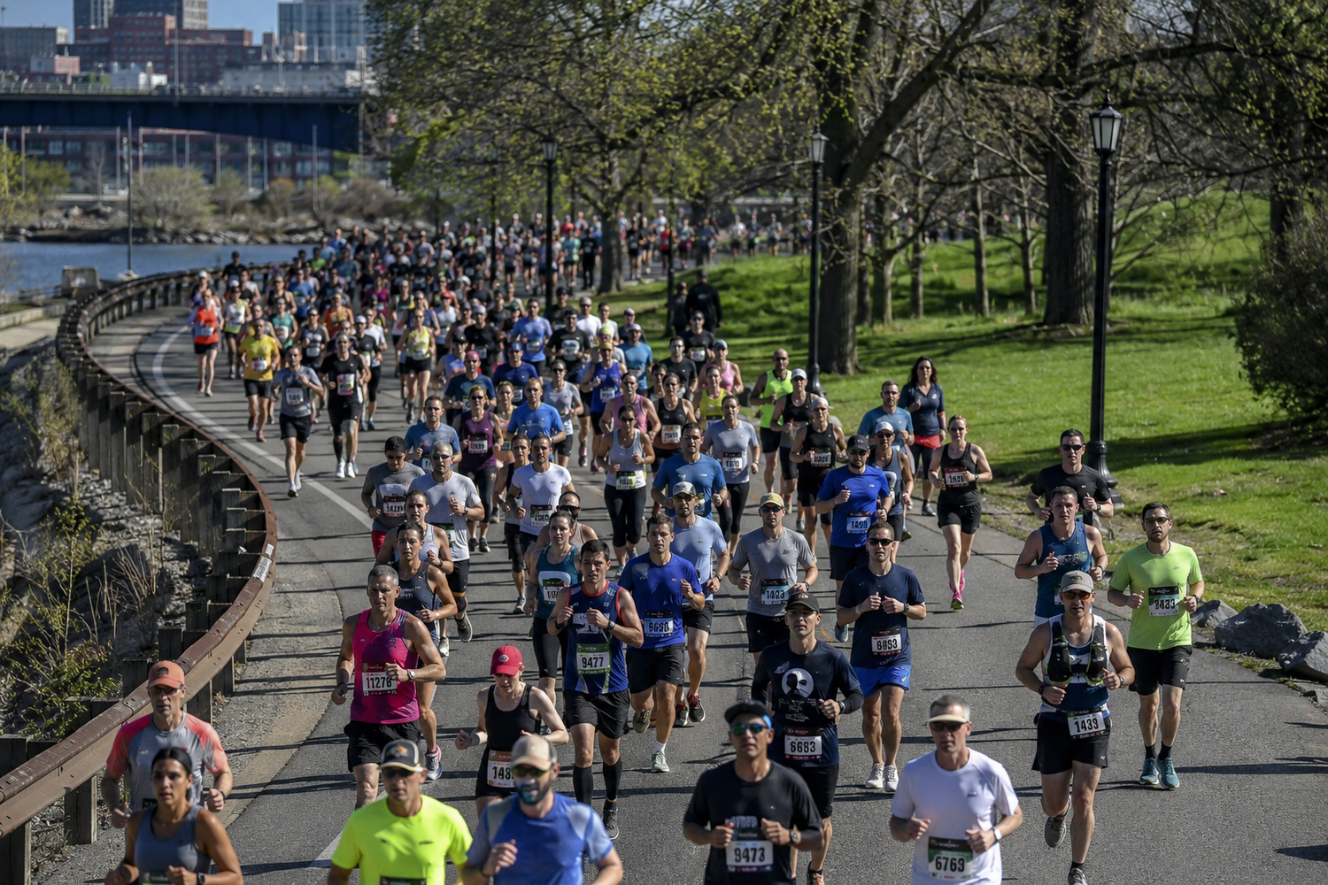 Large crowd of runners participating in a city marathon along a scenic riverside path on a sunny day.