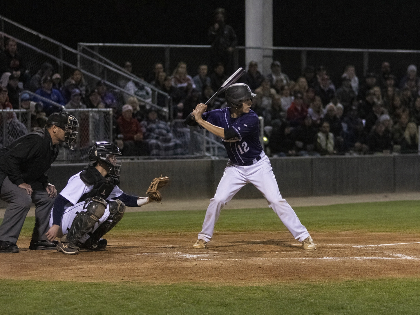 Baseball batter in purple jersey number 12 stands at home plate during a night game with catcher and umpire.