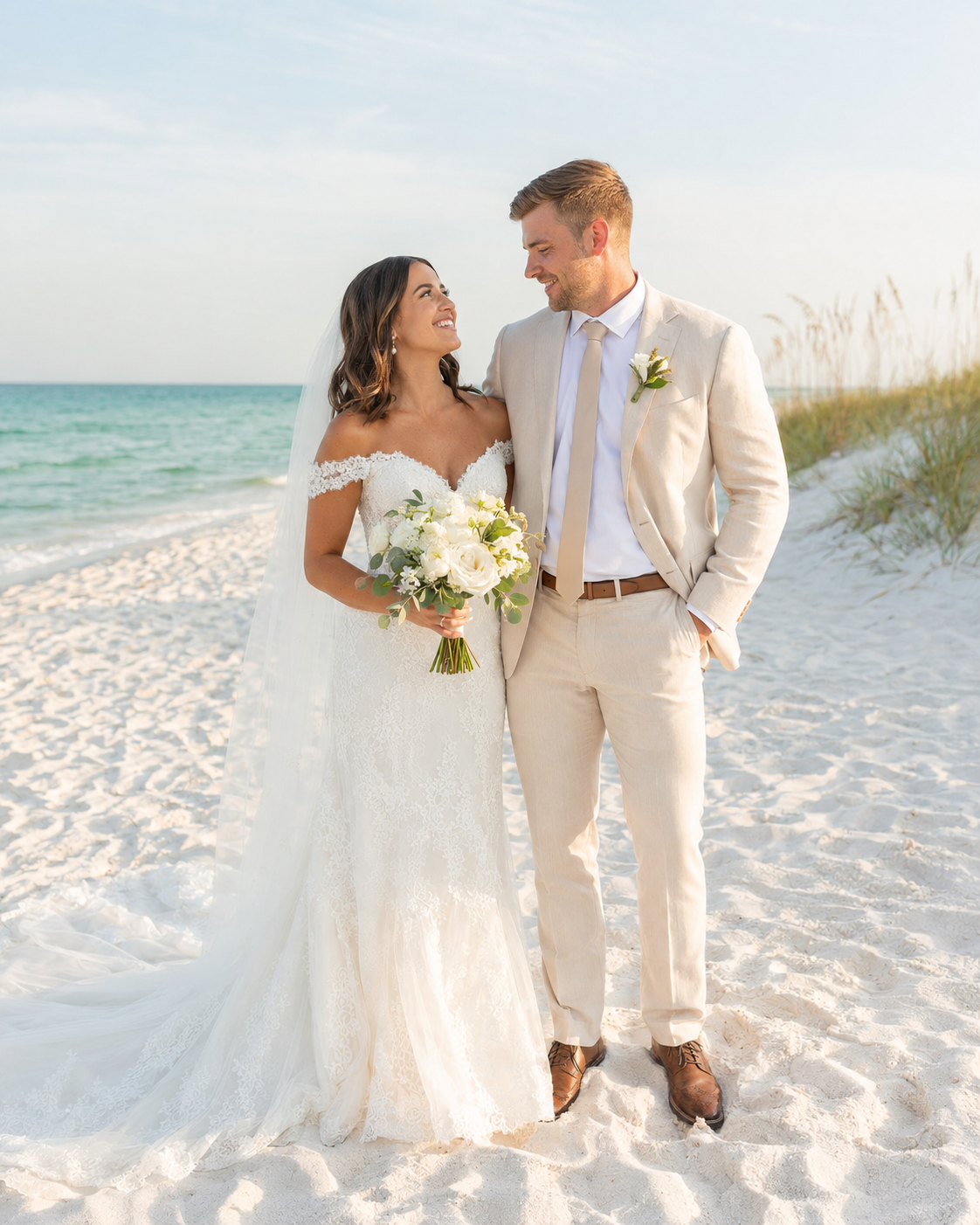 Bride and groom share a romantic moment on a white sand beach, she holds a white bouquet, he wears a tan suit.