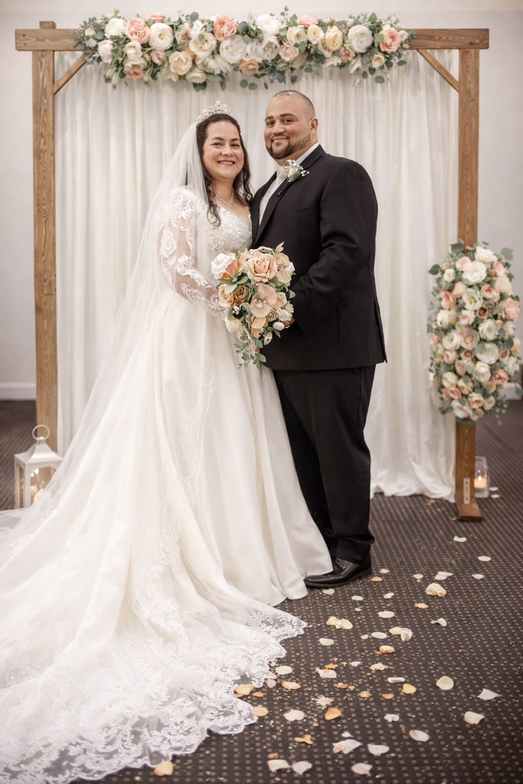Bride in white lace gown and groom in black tuxedo pose before floral arch with rose petals on floor.
