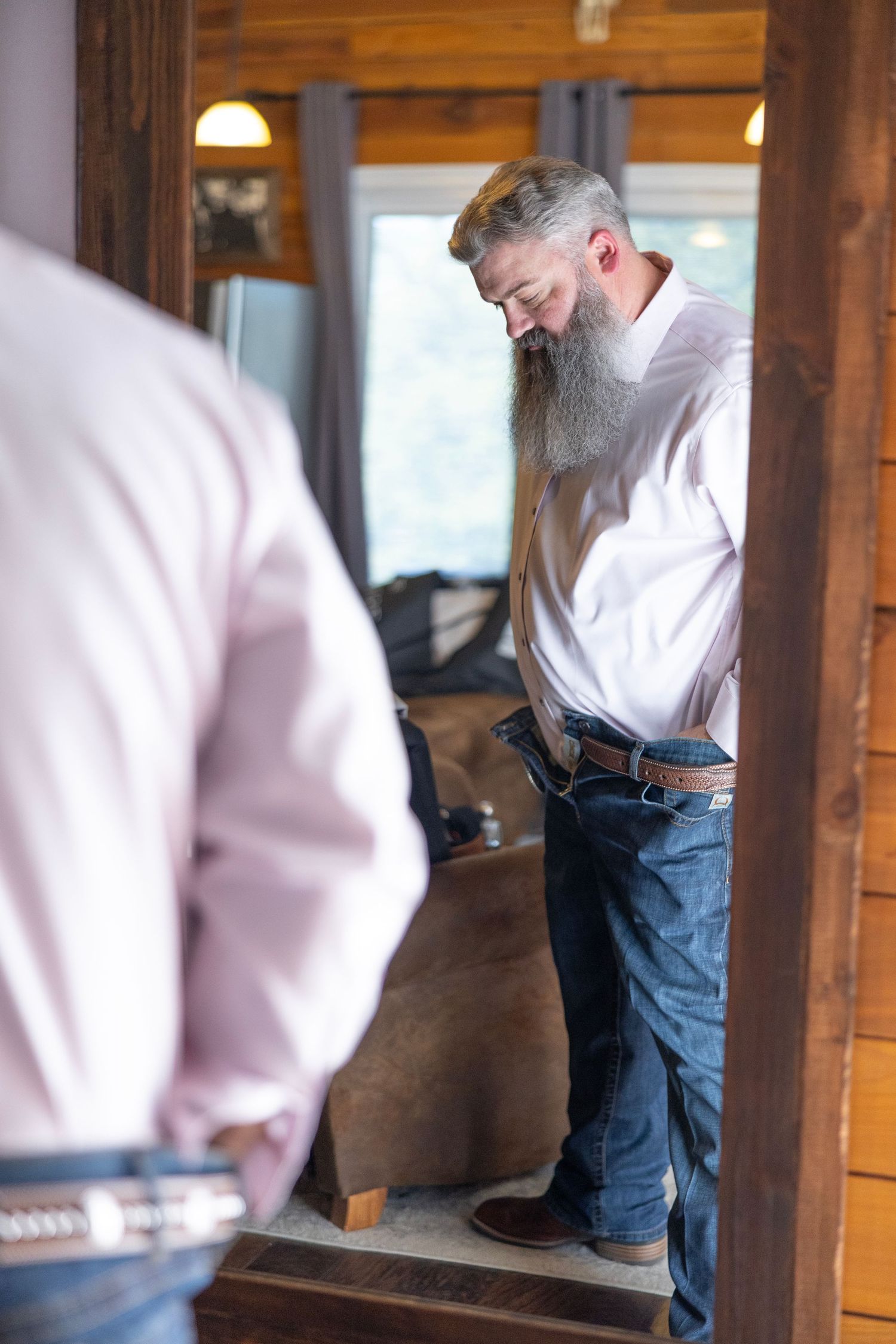 Bearded man in pink shirt and jeans checks his reflection in a rustic wooden mirror indoors.