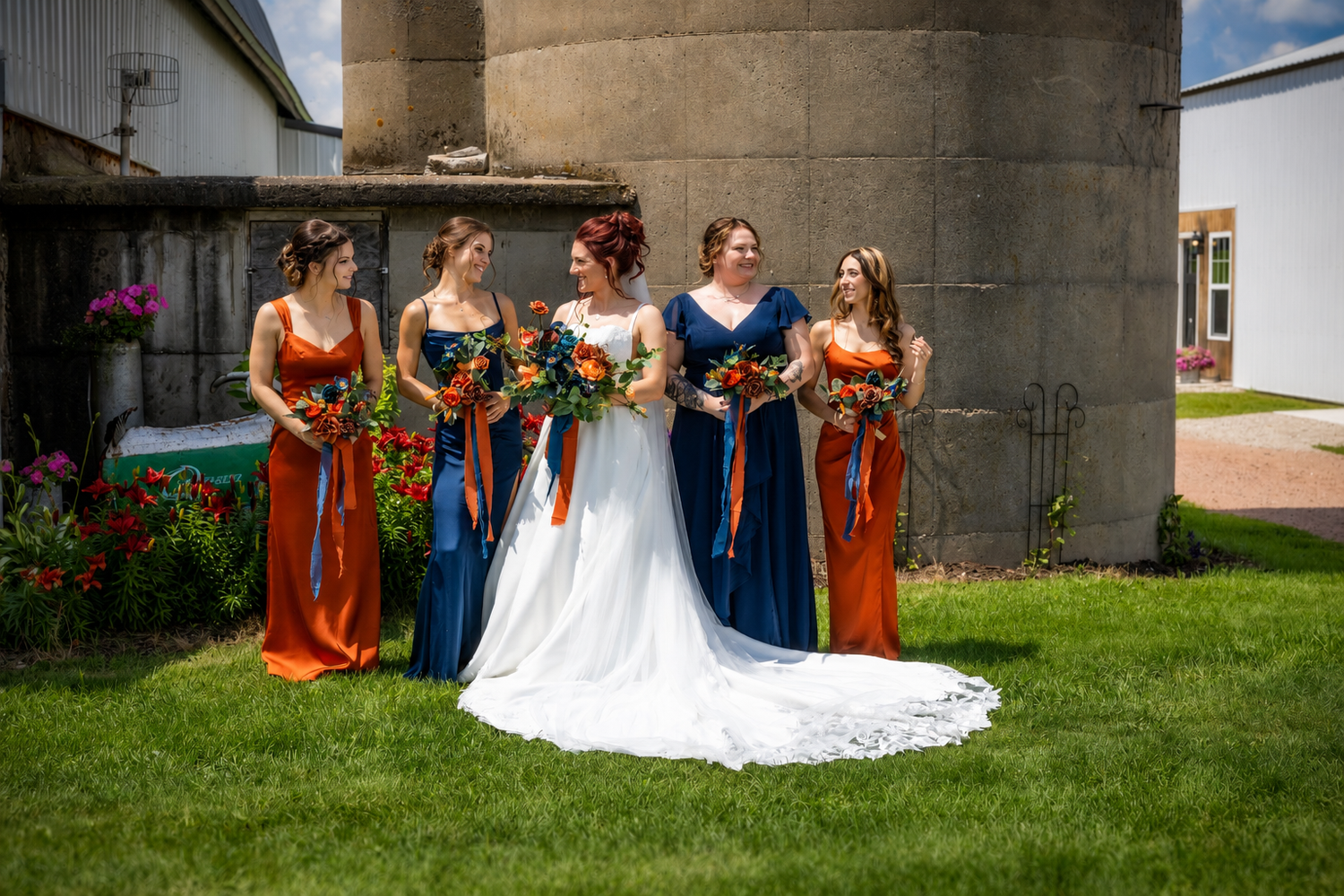 Bride in white gown with bridesmaids in navy and burnt orange dresses holding bouquets outdoors near a farm silo.