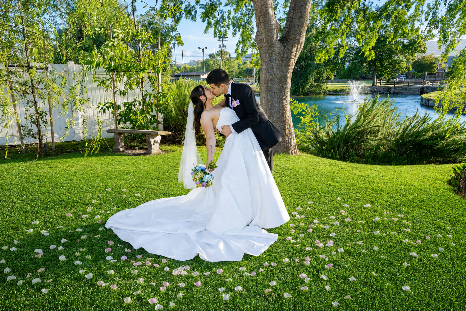 Bride and groom share a romantic dip kiss on lush green lawn with white flower petals scattered around them.
