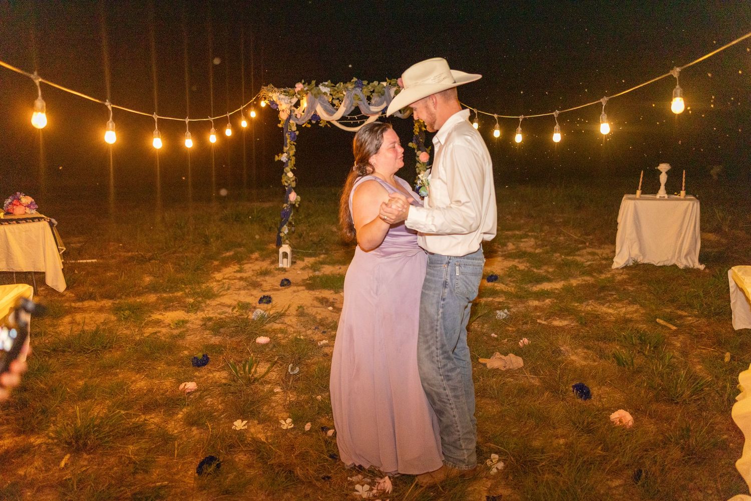 Couple sharing first dance outdoors at night under string lights at a rustic country wedding reception.