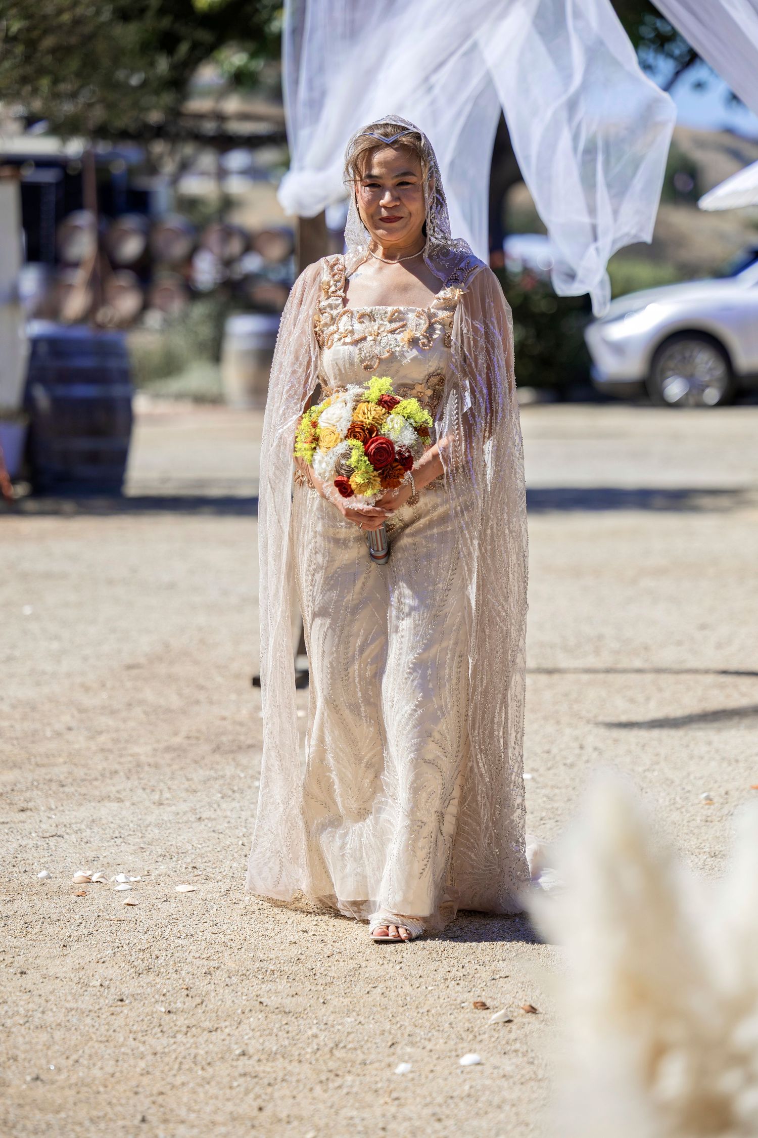 Bride in lace wedding jumpsuit and veil walks down sandy aisle holding colorful bouquet at outdoor ceremony.
