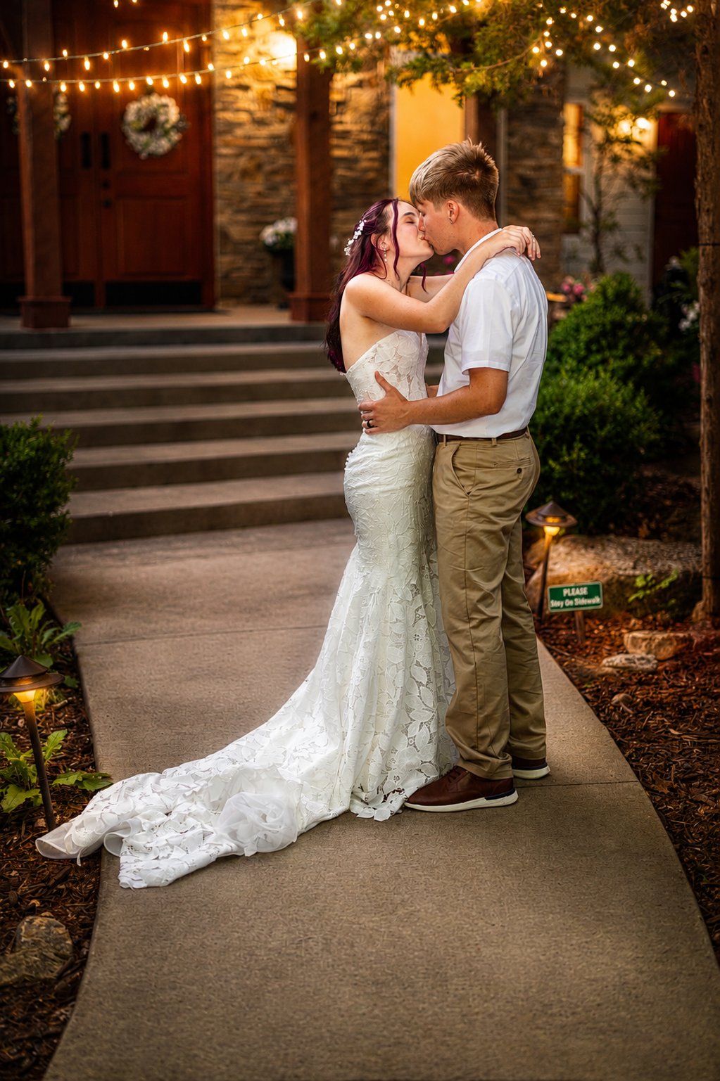 Bride and groom share a romantic kiss on a garden pathway at dusk, surrounded by warm string lights.