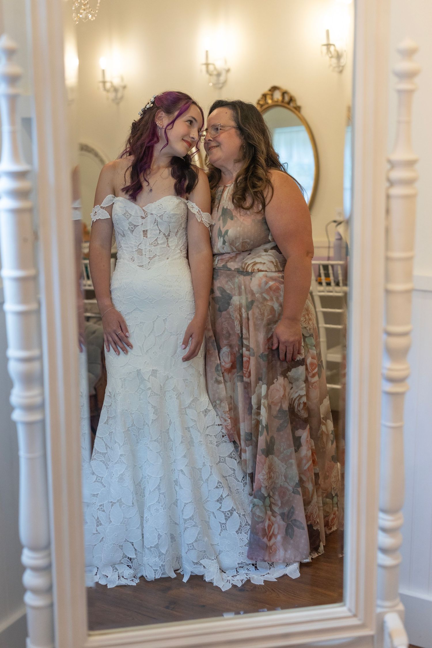 Bride in white lace mermaid gown and guest in floral dress share a joyful moment reflected in an ornate mirror.