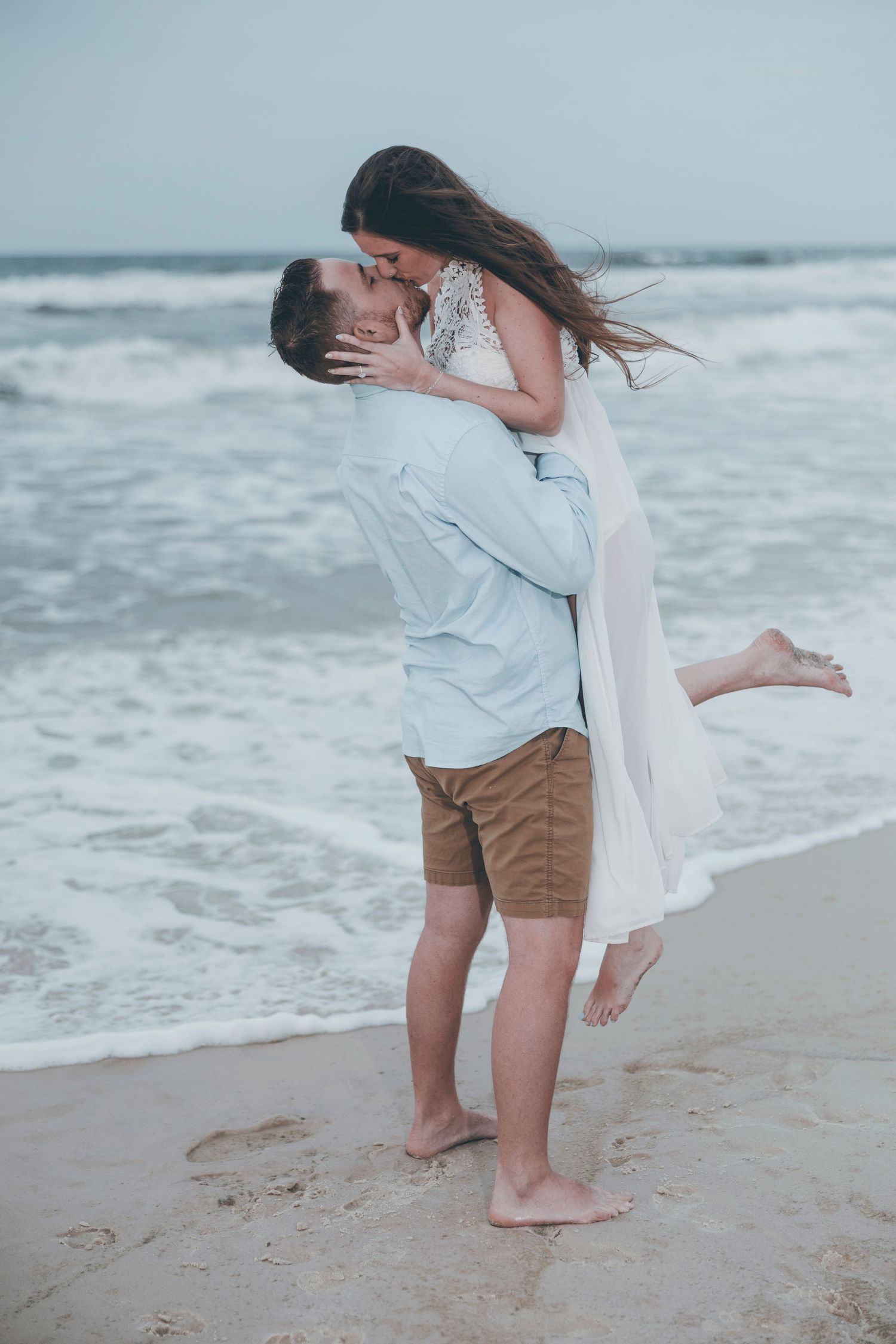 A romantic couple kissing on a sandy beach, man lifting woman in white dress, ocean waves in background.