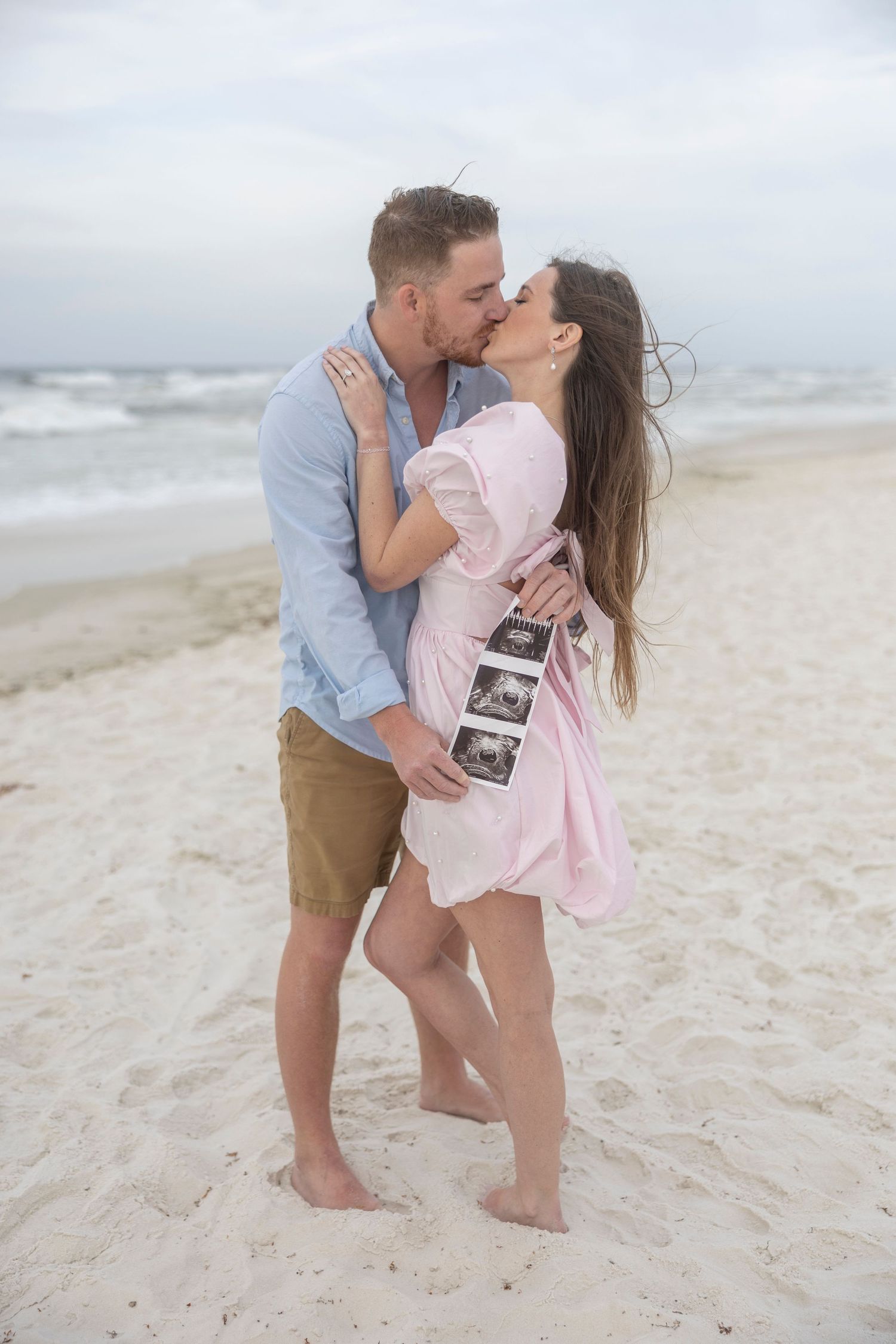 Couple kissing on beach holding ultrasound photos, pregnancy announcement on sandy shore with ocean waves in background.