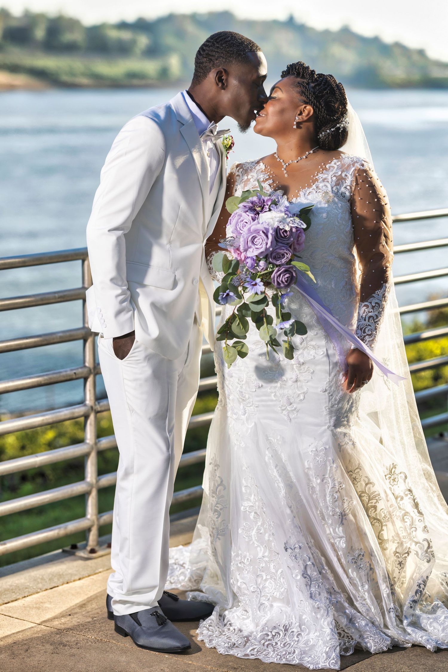 A Black bride and groom share a romantic moment in wedding attire, bride holding purple roses bouquet near waterfront.