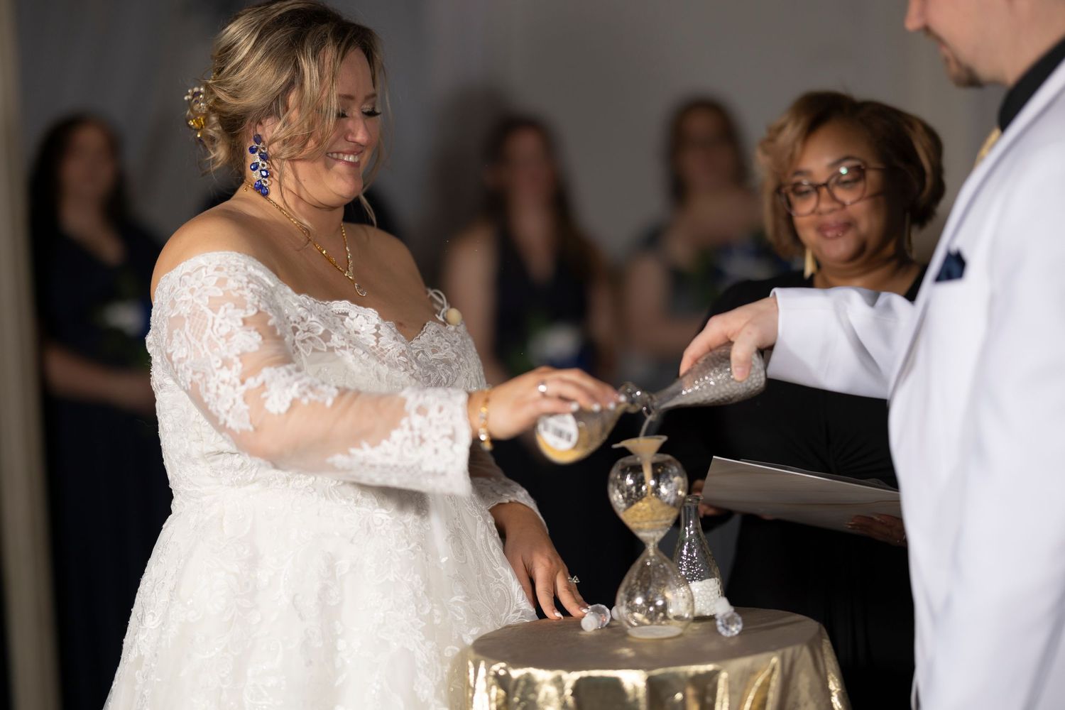 Bride in lace wedding gown performs sand ceremony at wedding reception with guests in navy dresses in background.