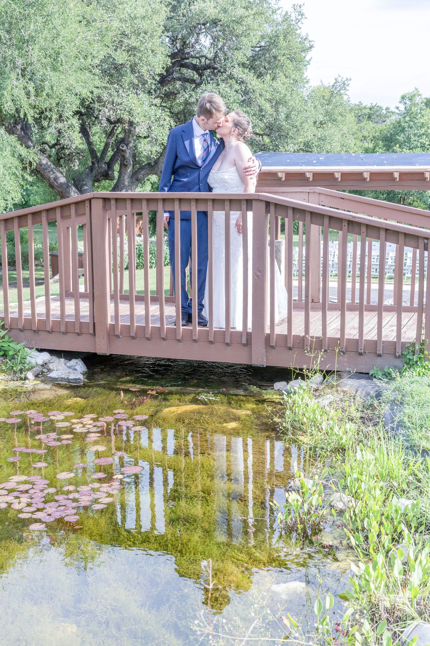 A couple stands together on a wooden footbridge over a lily pond surrounded by lush greenery on a sunny day.