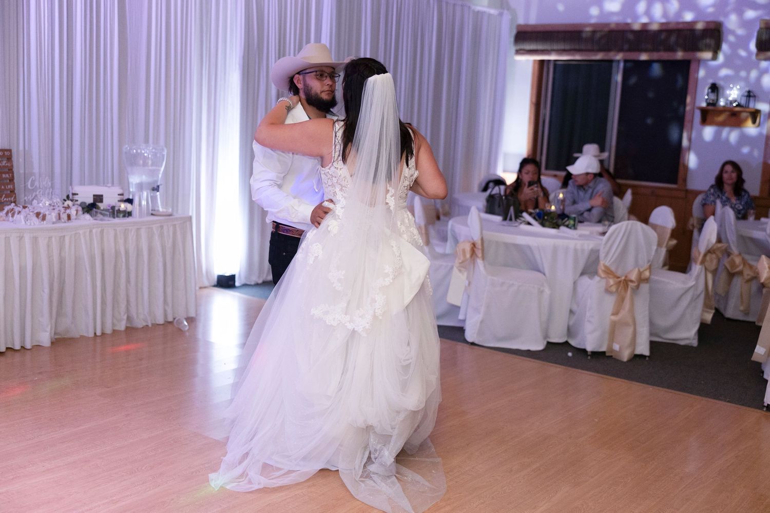 A couple shares their first dance at a wedding reception with white decor and chair covers on a polished wood floor.