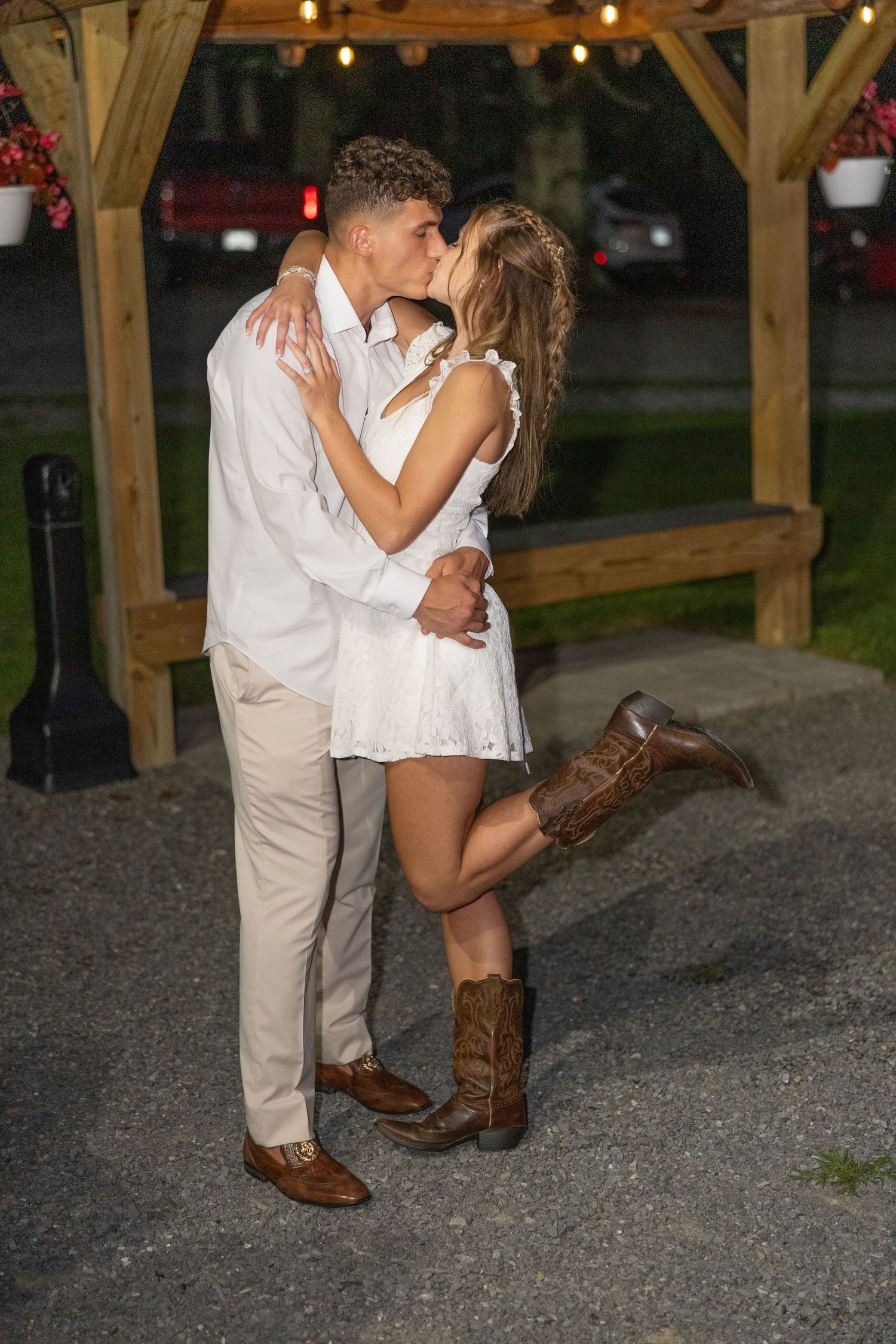 A romantic couple shares a kiss at dusk under a wooden gazebo while wearing cowboy boots and casual attire.