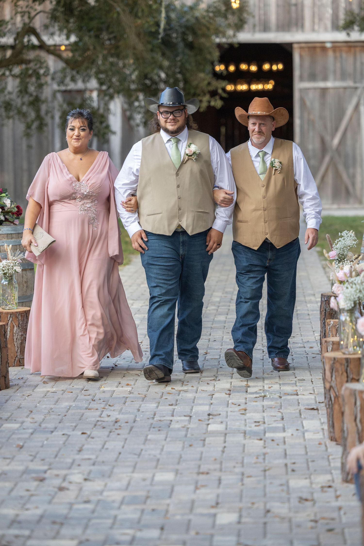 Wedding guests in country western attire walk down a rustic brick pathway at an outdoor barn venue.