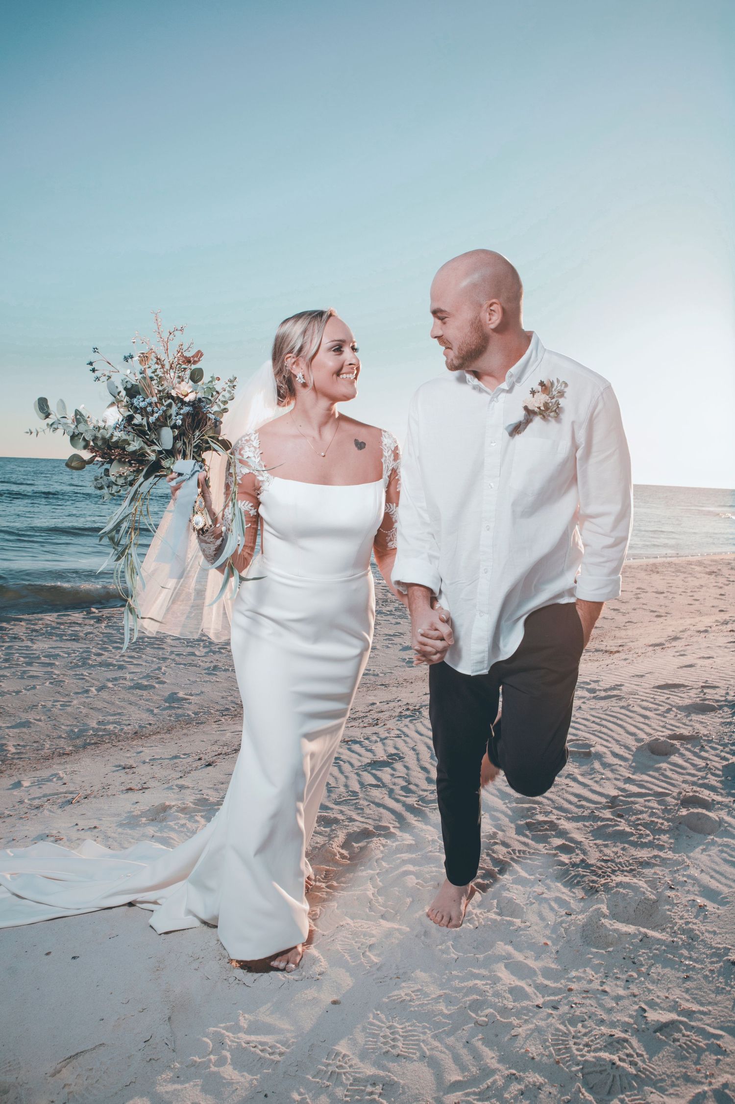 A couple walks barefoot along a sandy beach at sunset in wedding attire.