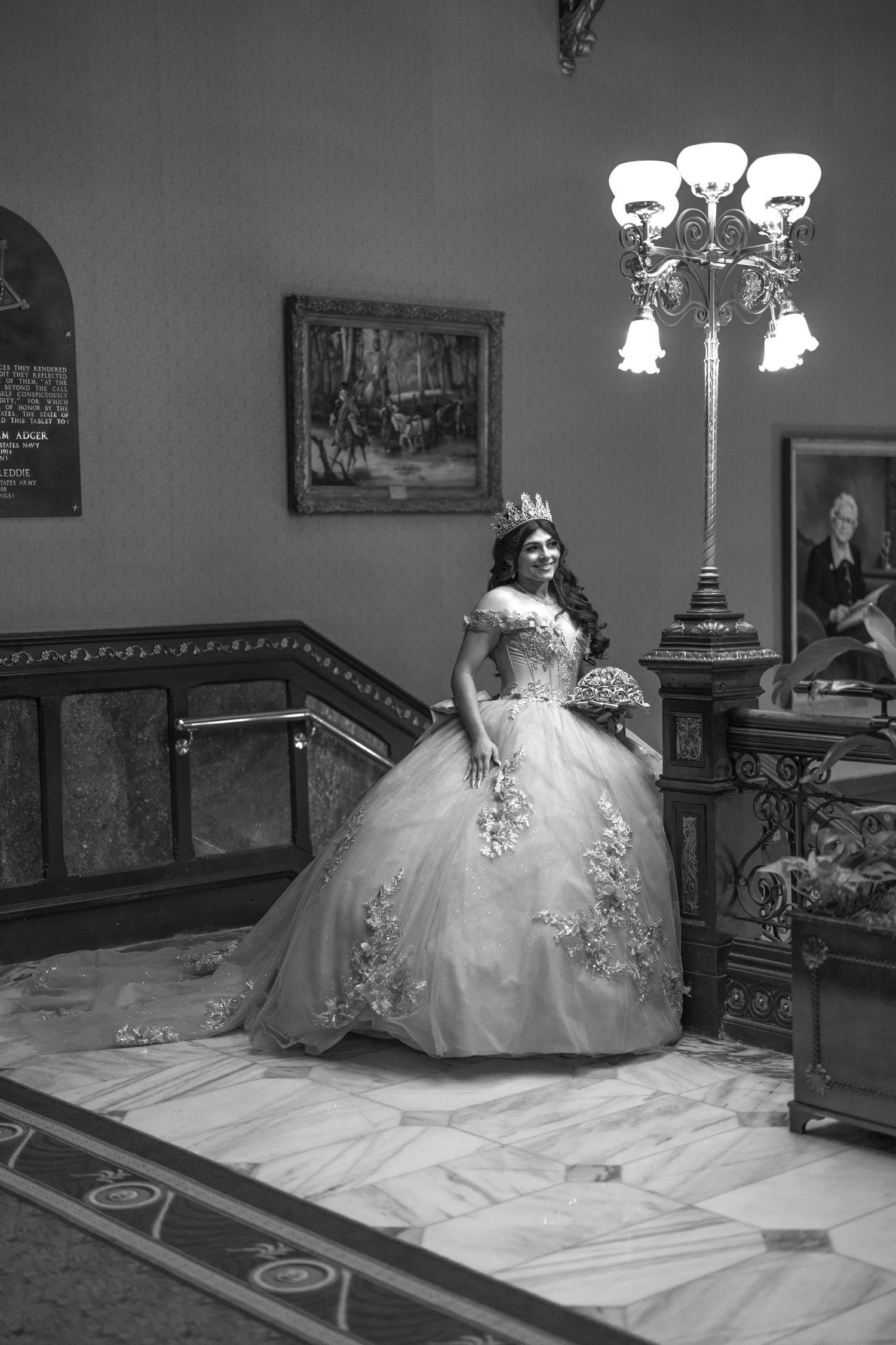 A bride in an elegant white ball gown with floral details poses on a marble floor near an ornate vintage lamp and staircase.