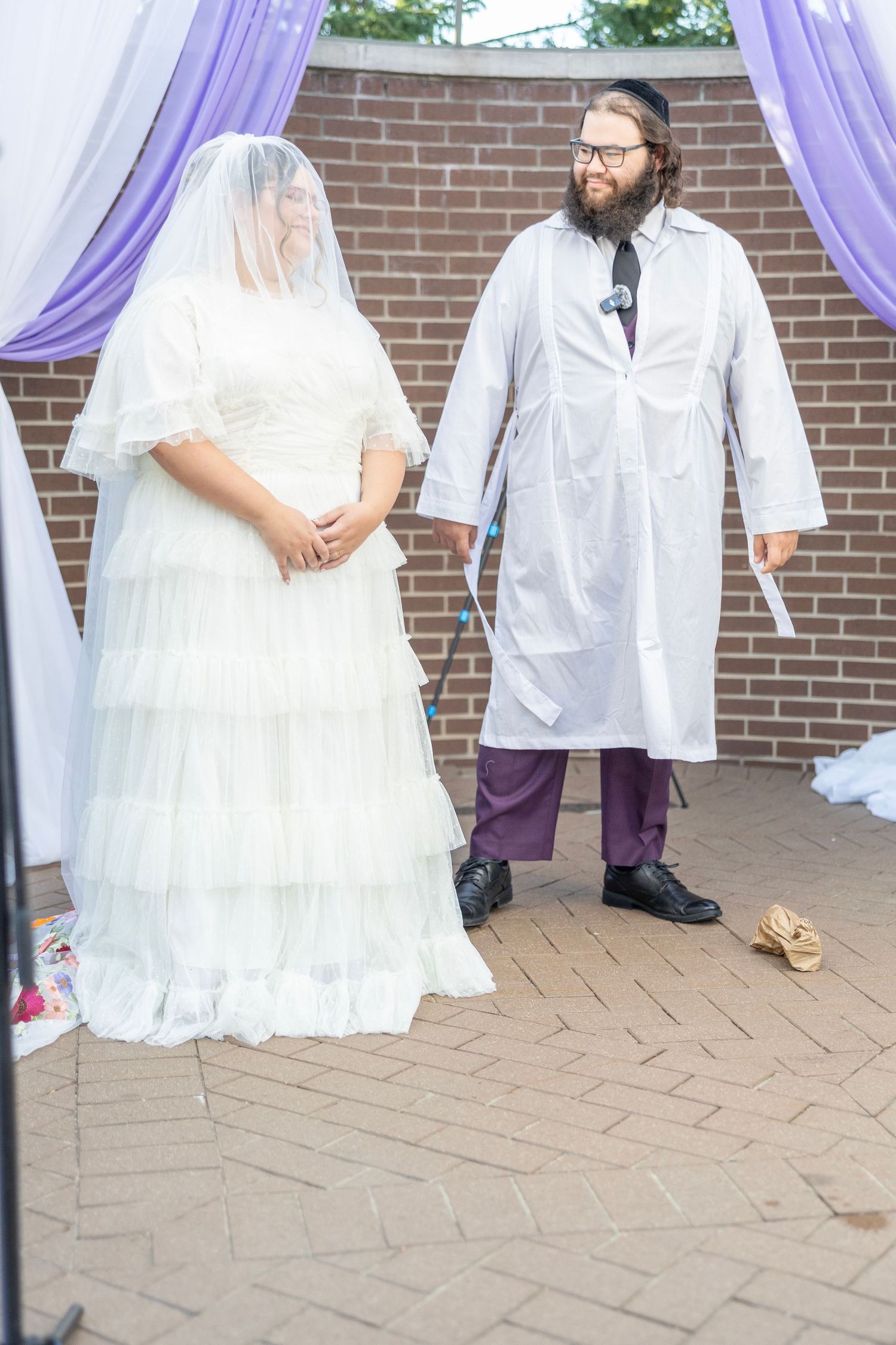 A wedding ceremony under purple drapes with a bride in a white lace gown and someone wearing a white lab coat.